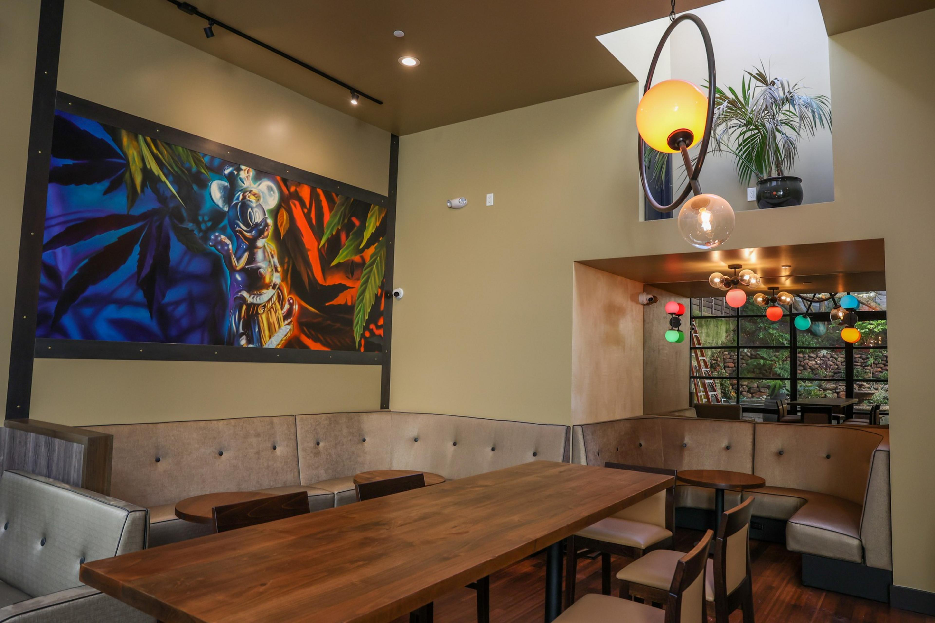 The image shows a modern dining area with a long wooden table, cushioned booth seating, a colorful mural, and hanging spherical lights. A plant is visible above.