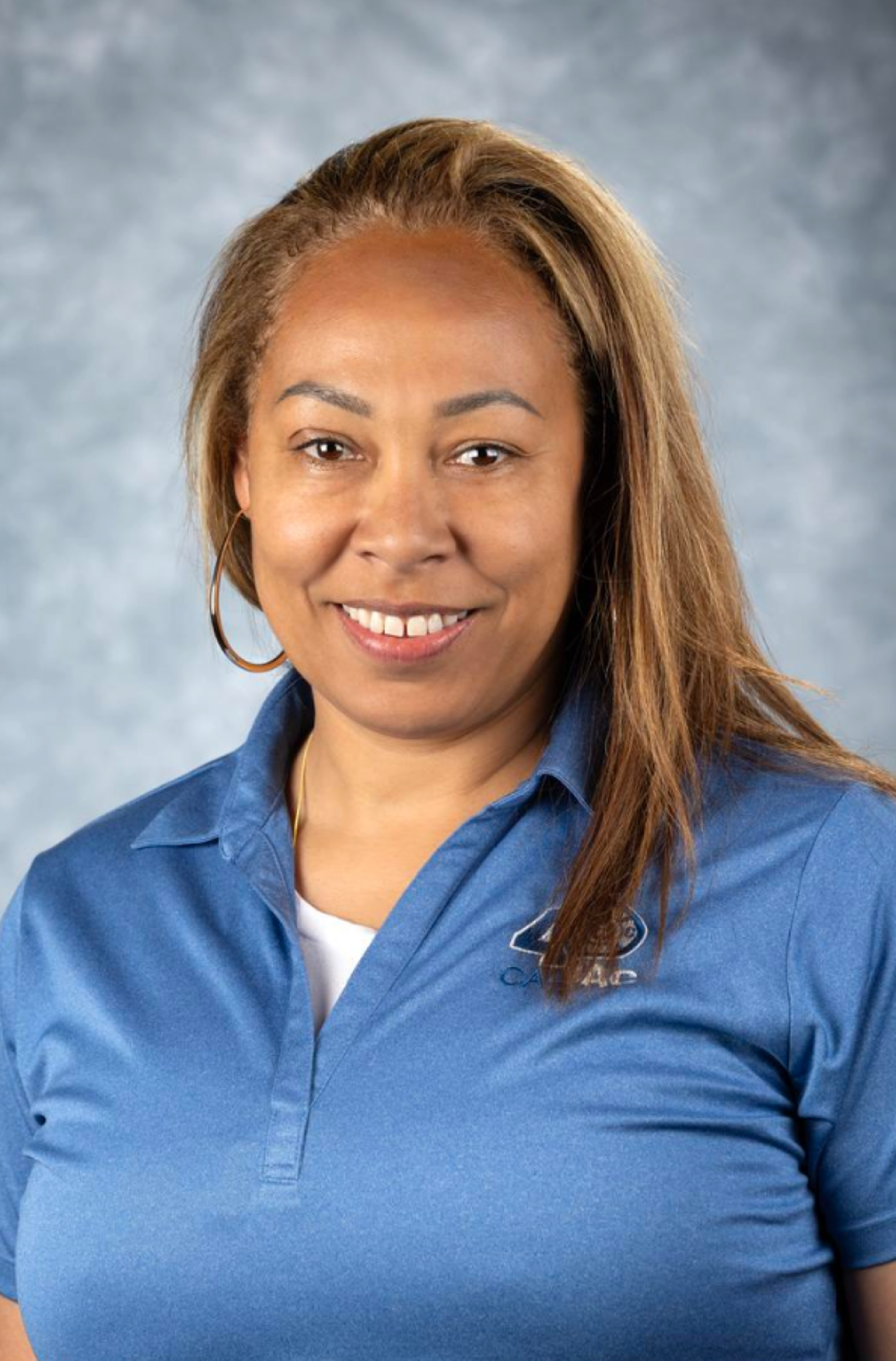 A woman with light brown hair, wearing large hoop earrings and a blue polo shirt with a logo, smiles against a soft gray background.