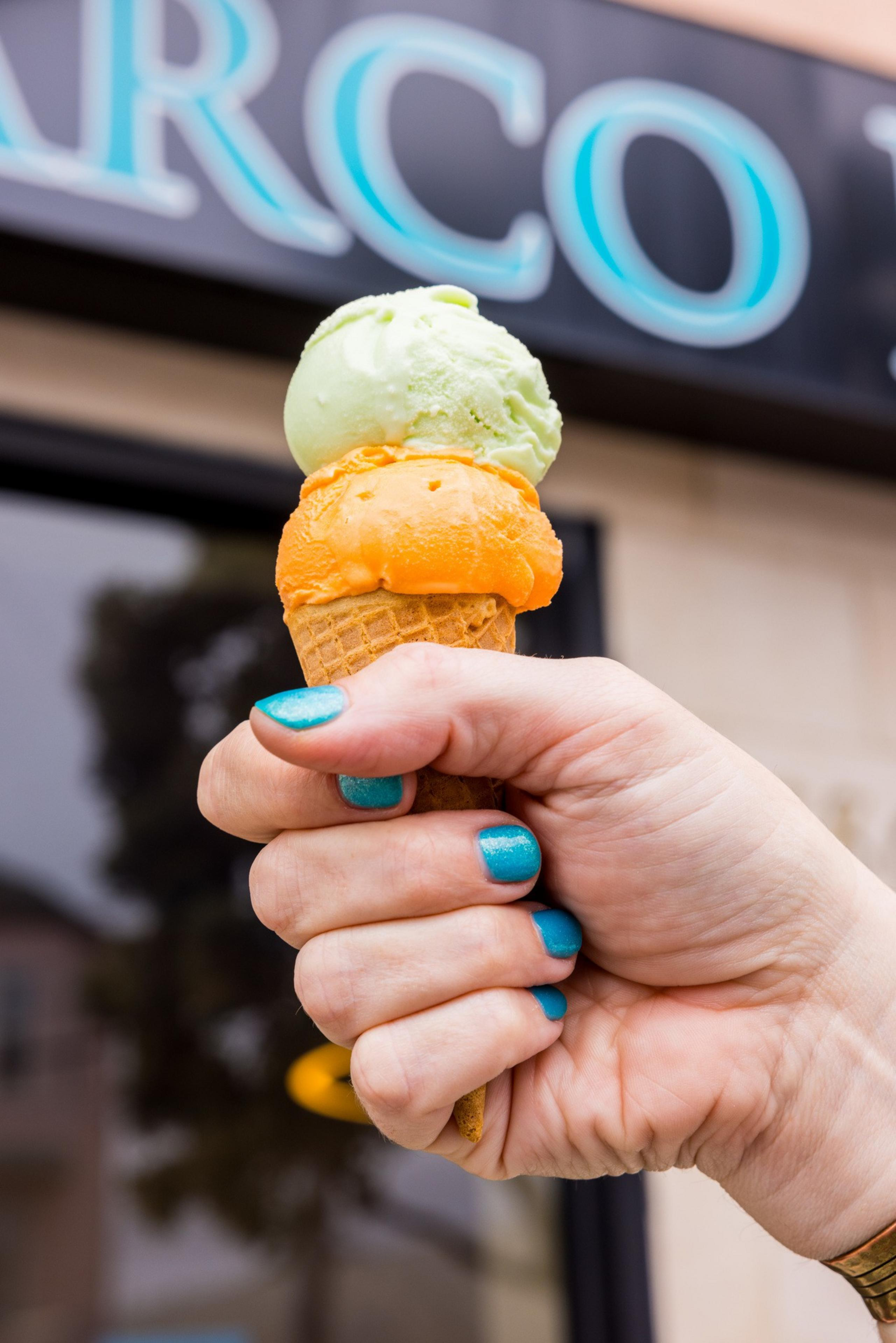 A hand with turquoise nails holds an ice cream cone with two scoops, one orange and one green, in front of a blurred background with partial signage.