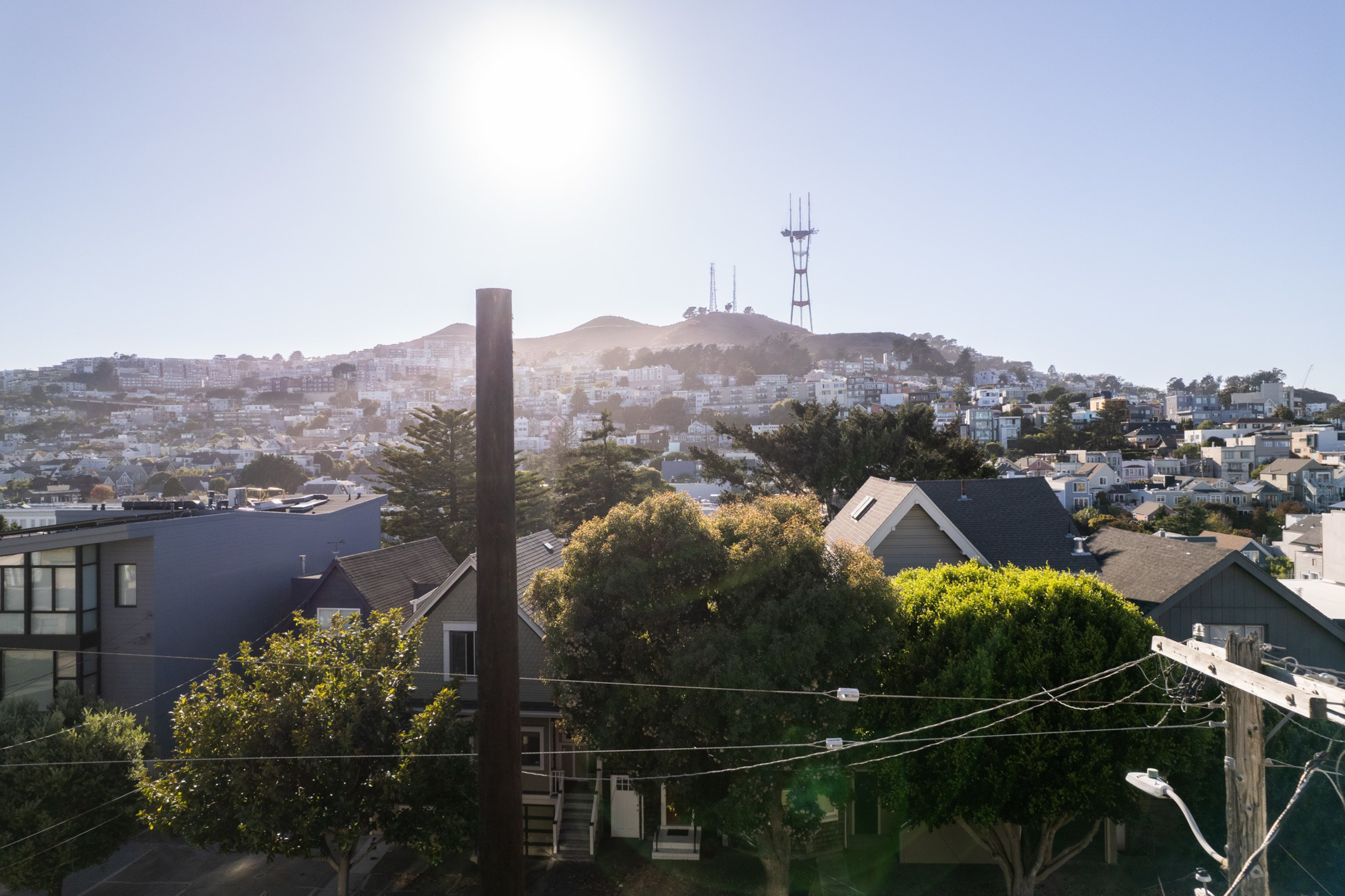 A sunny view of a residential area with trees, houses, and power lines in front, with a hill and a tall communication tower in the background.