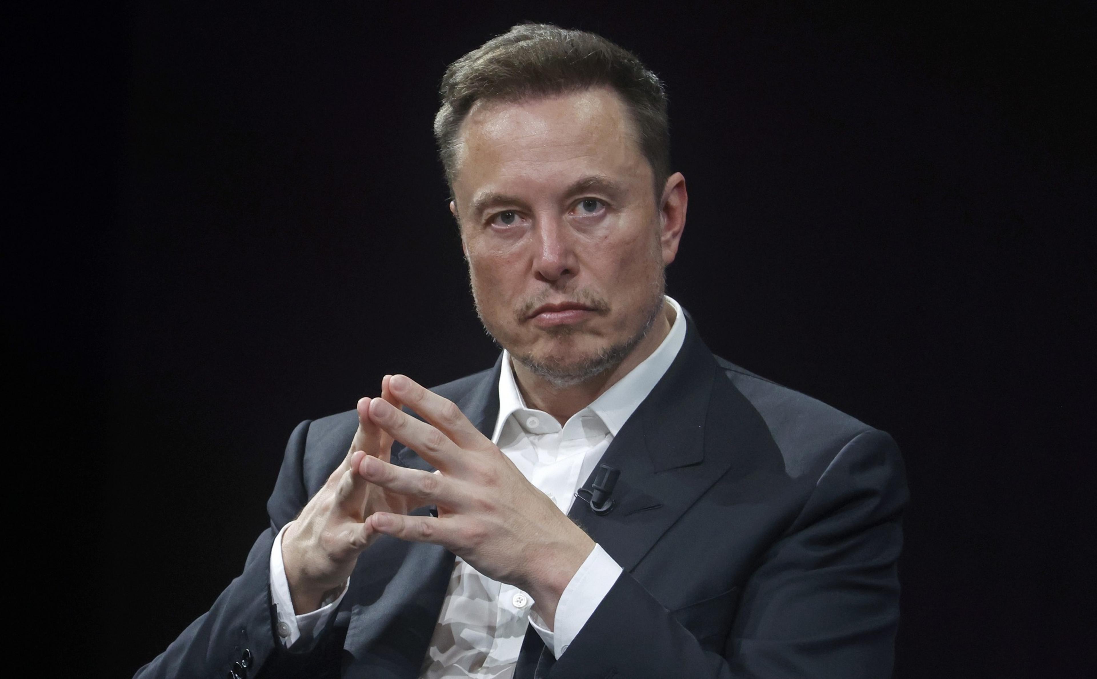A man with short, neat hair and light facial stubble is looking directly at the camera. He wears a white shirt and dark suit, with hands interlocked in front of his face.