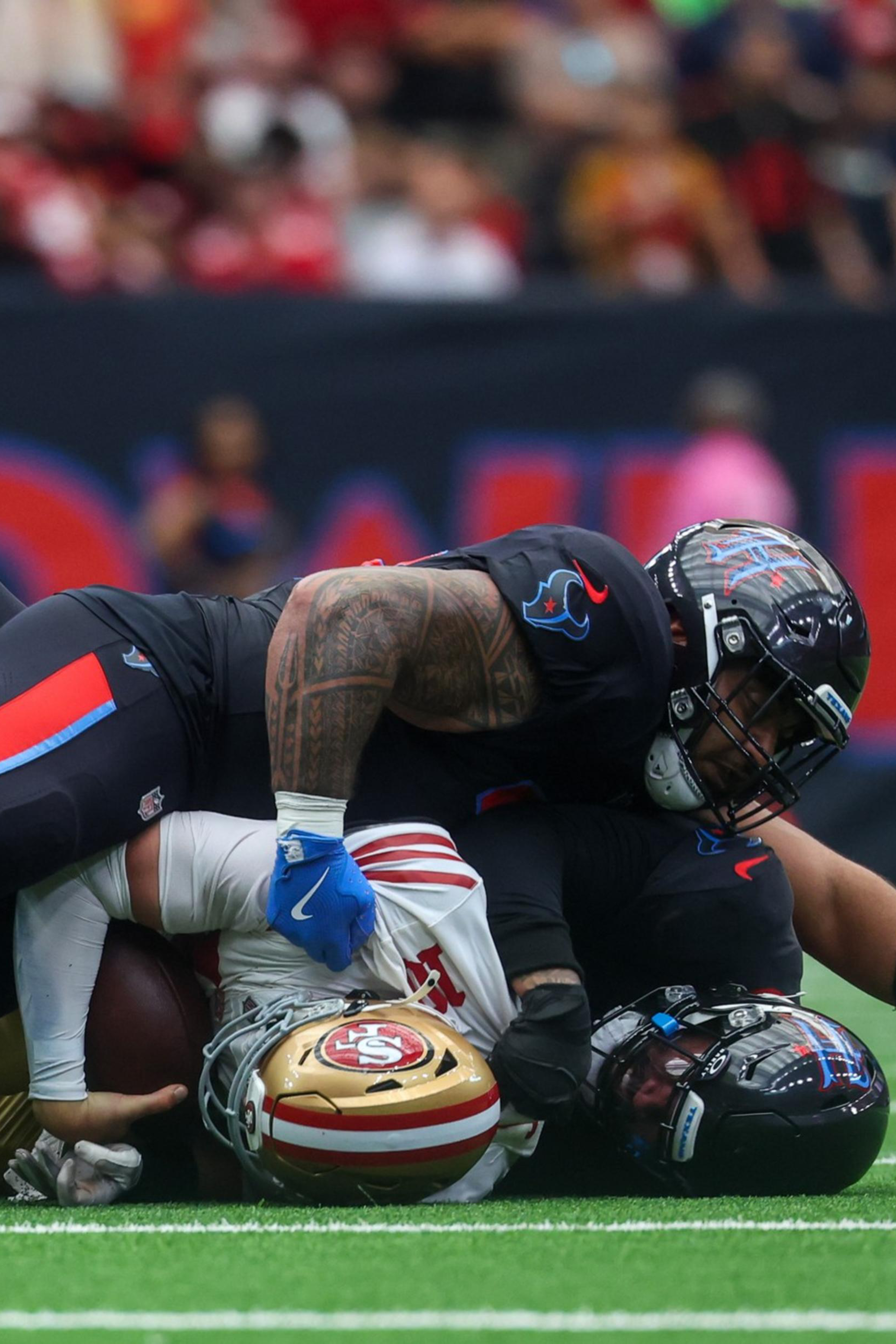 Two football players in black uniforms tackle a player in a white and red uniform who is holding the ball, all lying on a green field.