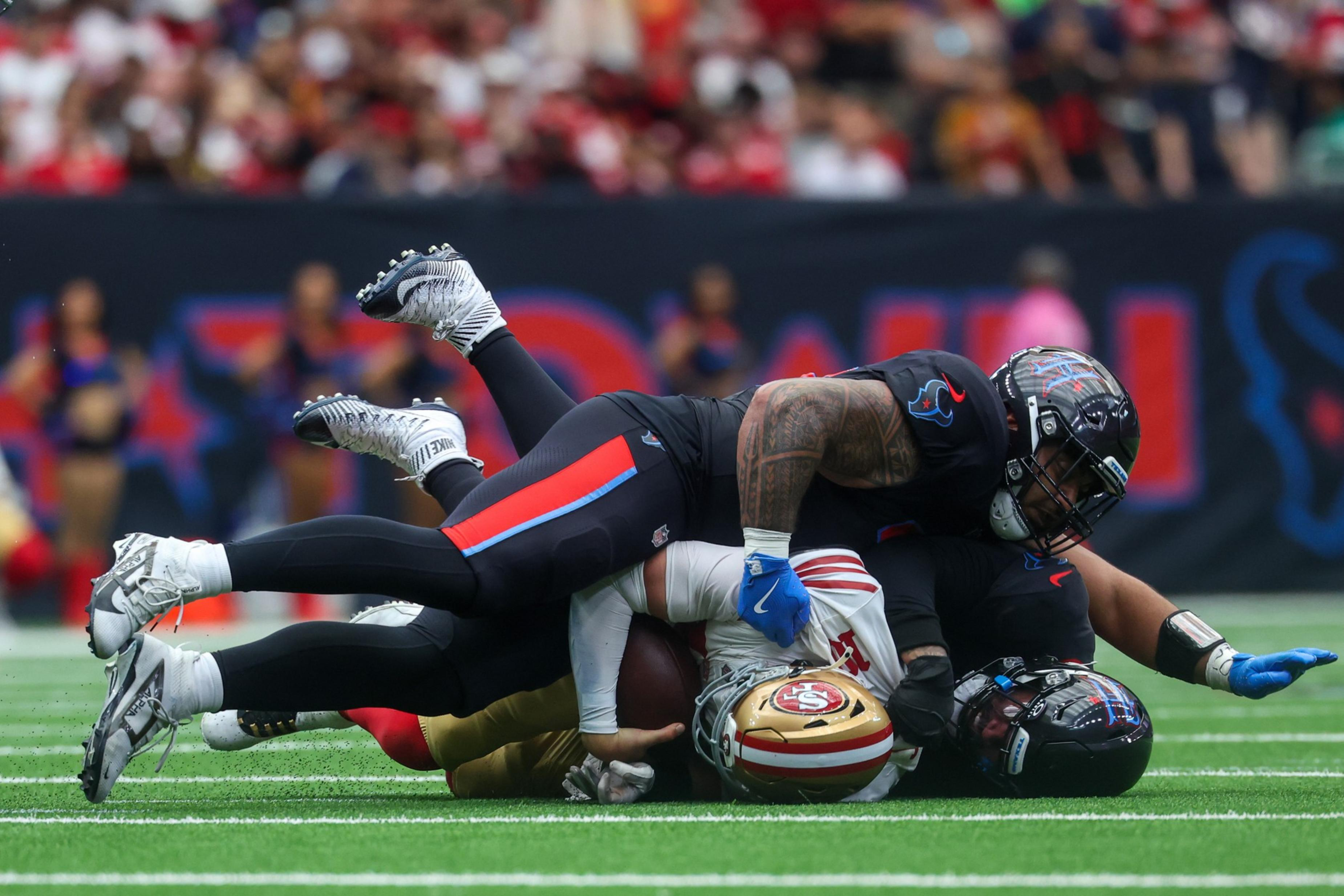 Two football players in black uniforms tackle a player in a white and red uniform who is holding the ball, all lying on a green field.