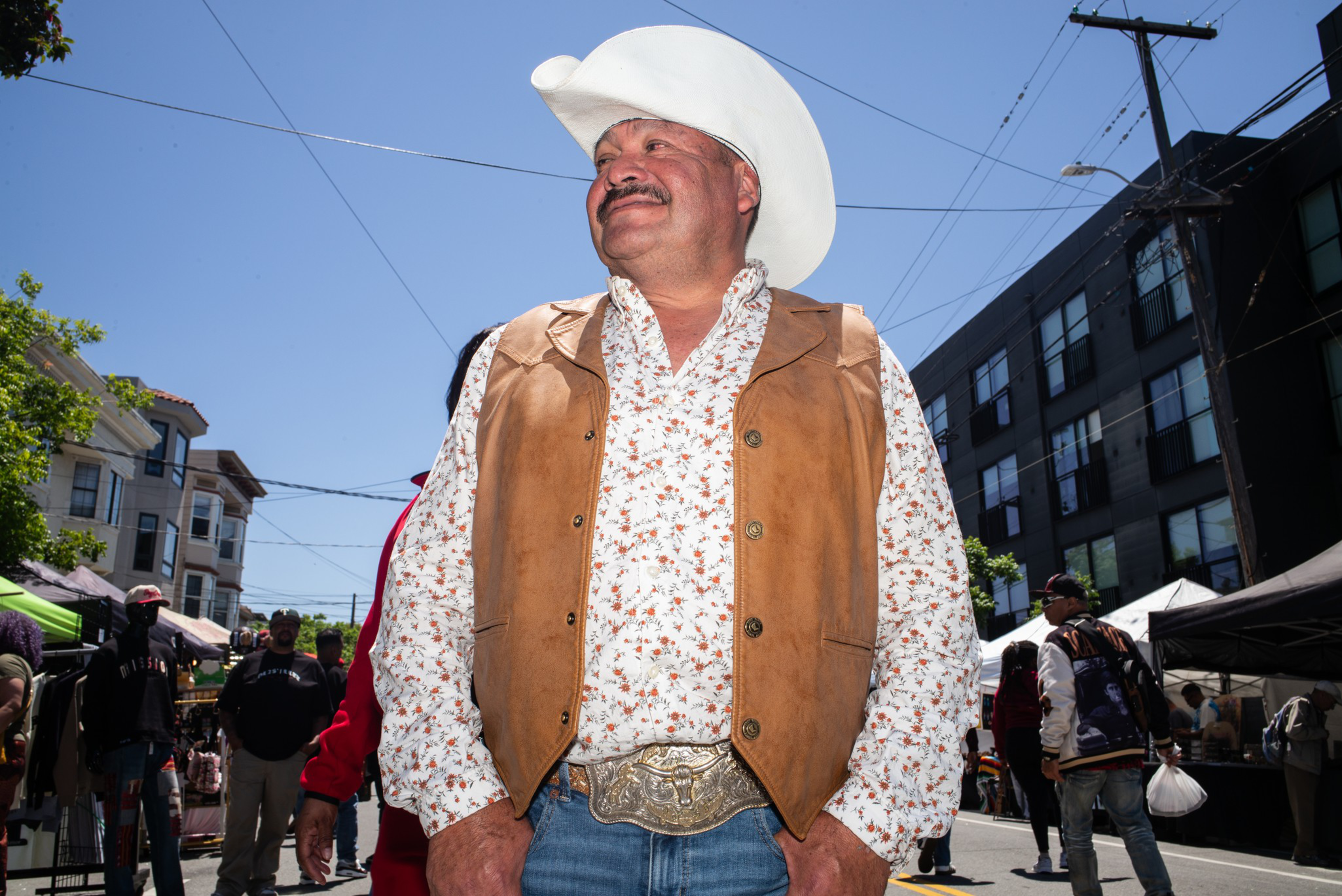 A man wearing a white cowboy hat, brown leather vest, floral shirt, and large belt buckle stands confidently outdoors on a sunny day with buildings and people behind him.