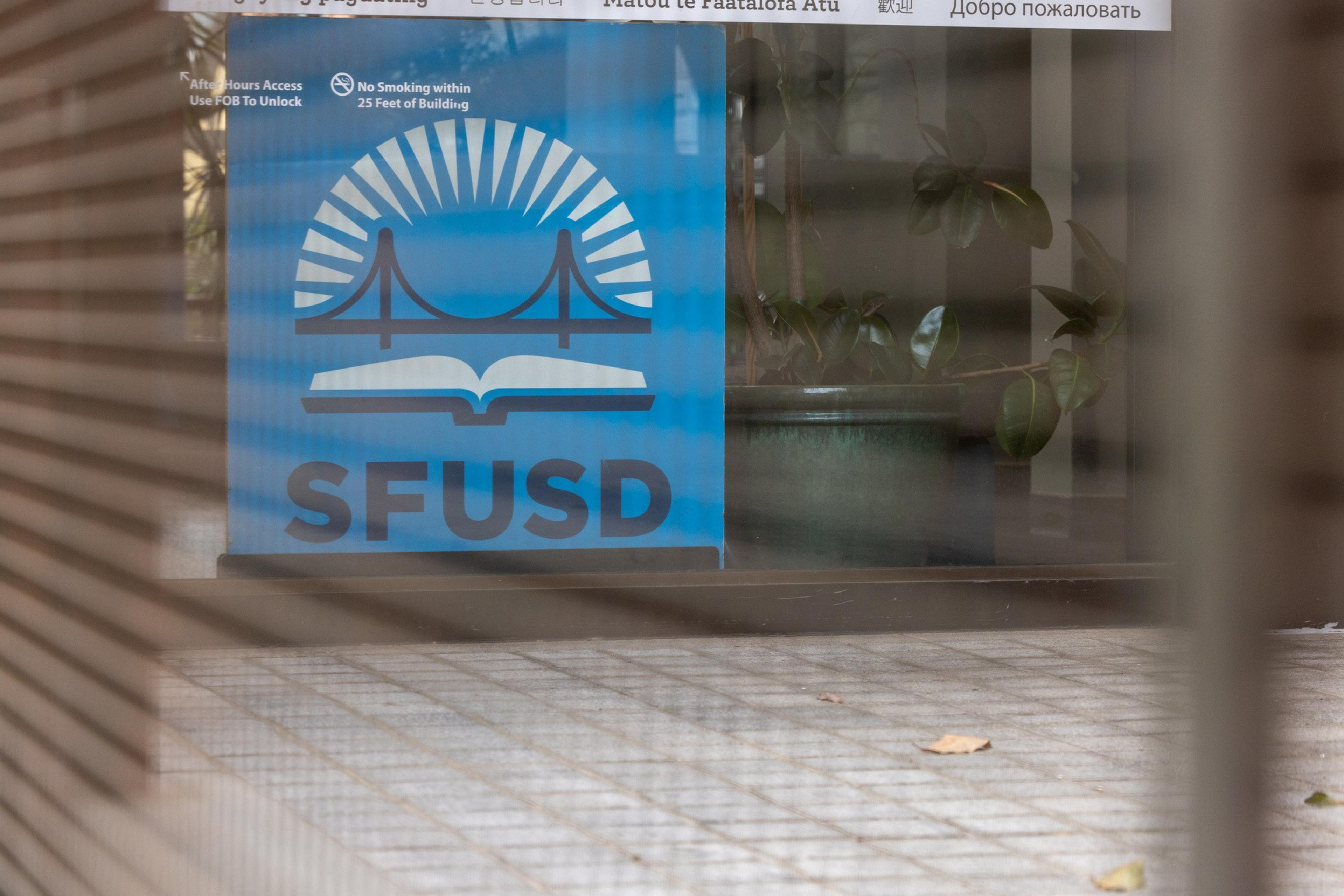 The image shows a sign with a blue background featuring a bridge and book logo, and the letters &quot;SFUSD.&quot; It's viewed through slats with a plant nearby.