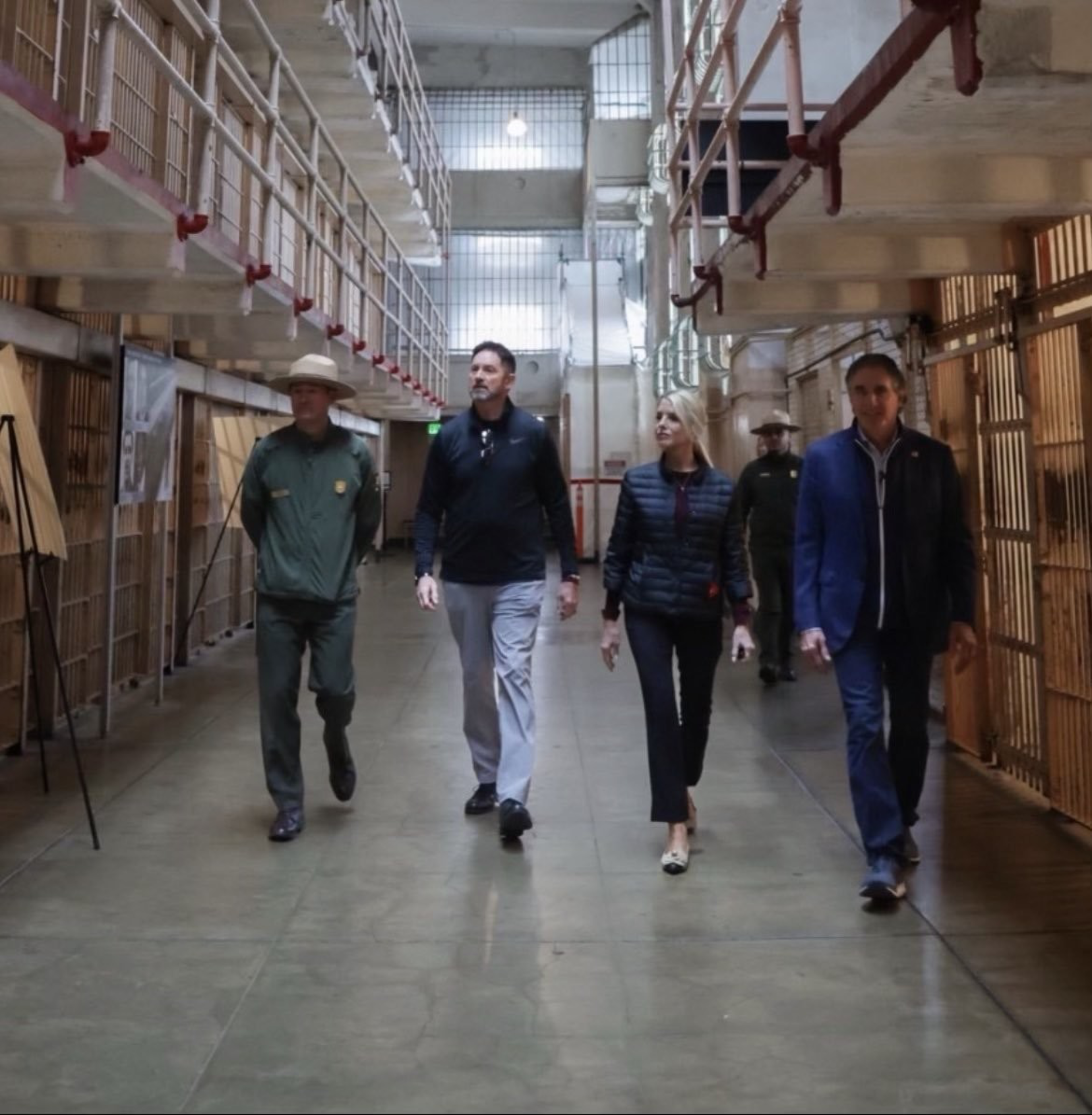 Four people are walking down a corridor in a prison, with metal bars and cell doors on either side. One person is in a green uniform.