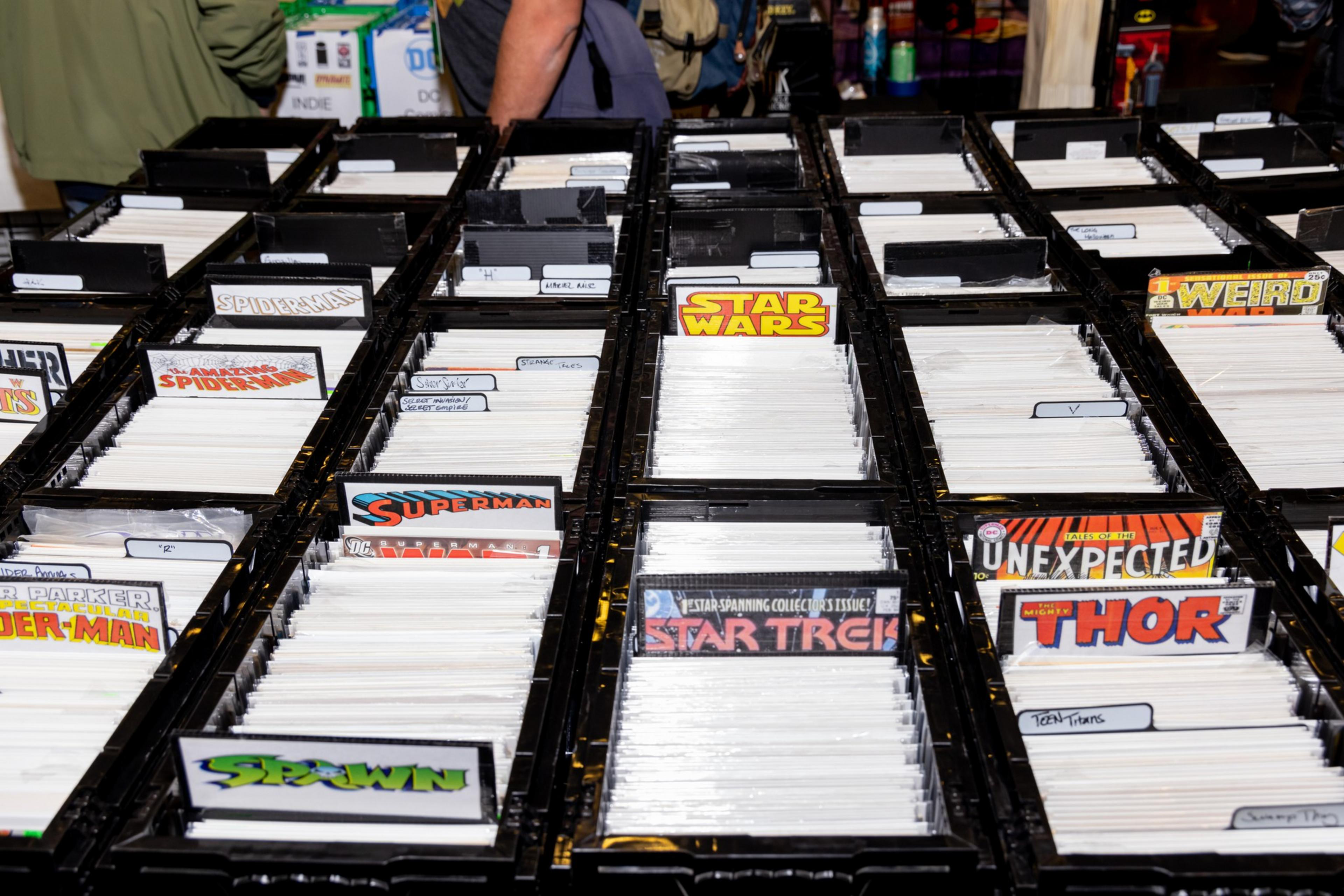Rows of black crates hold comic books with visible titles like Spider-Man, Star Wars, Superman, Star Trek, and Thor. People are browsing on the left.