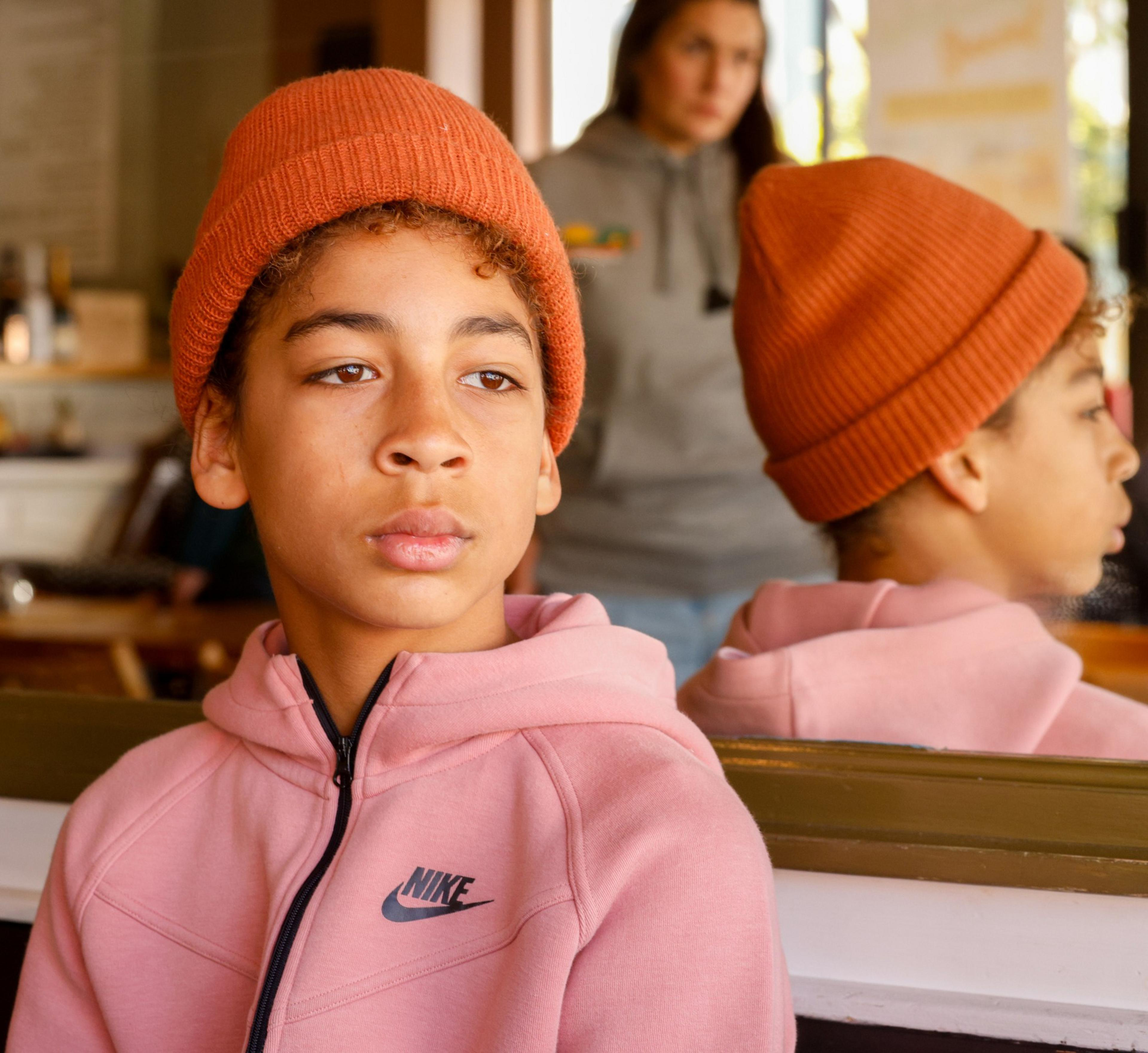 A young person in a pink hoodie and orange beanie, with a reflective expression, mirrored in the glass next to a distracted woman.