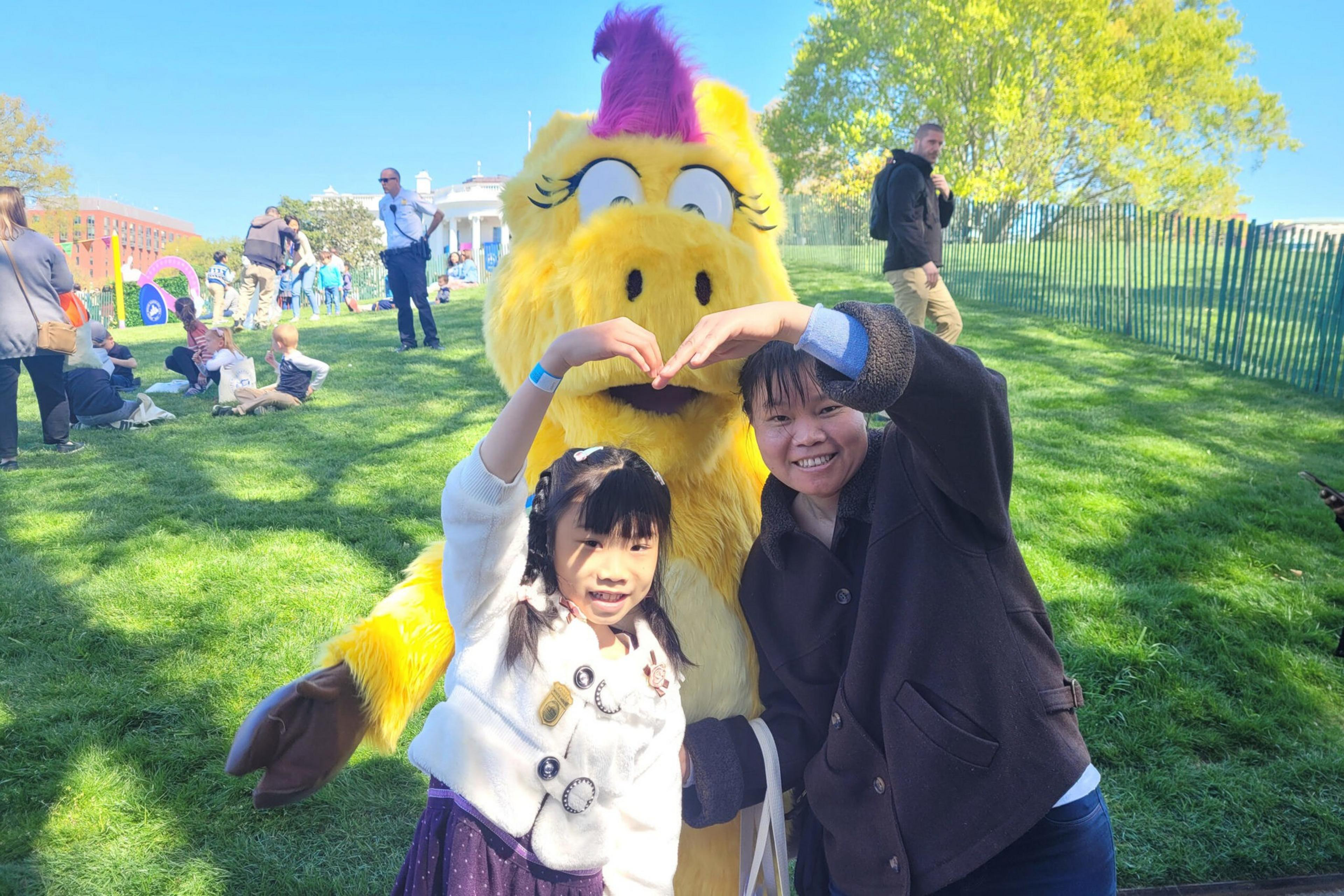 A child and an adult form a heart shape with their hands in front of a yellow bird mascot with purple and pink feathers on a sunny grassy lawn.