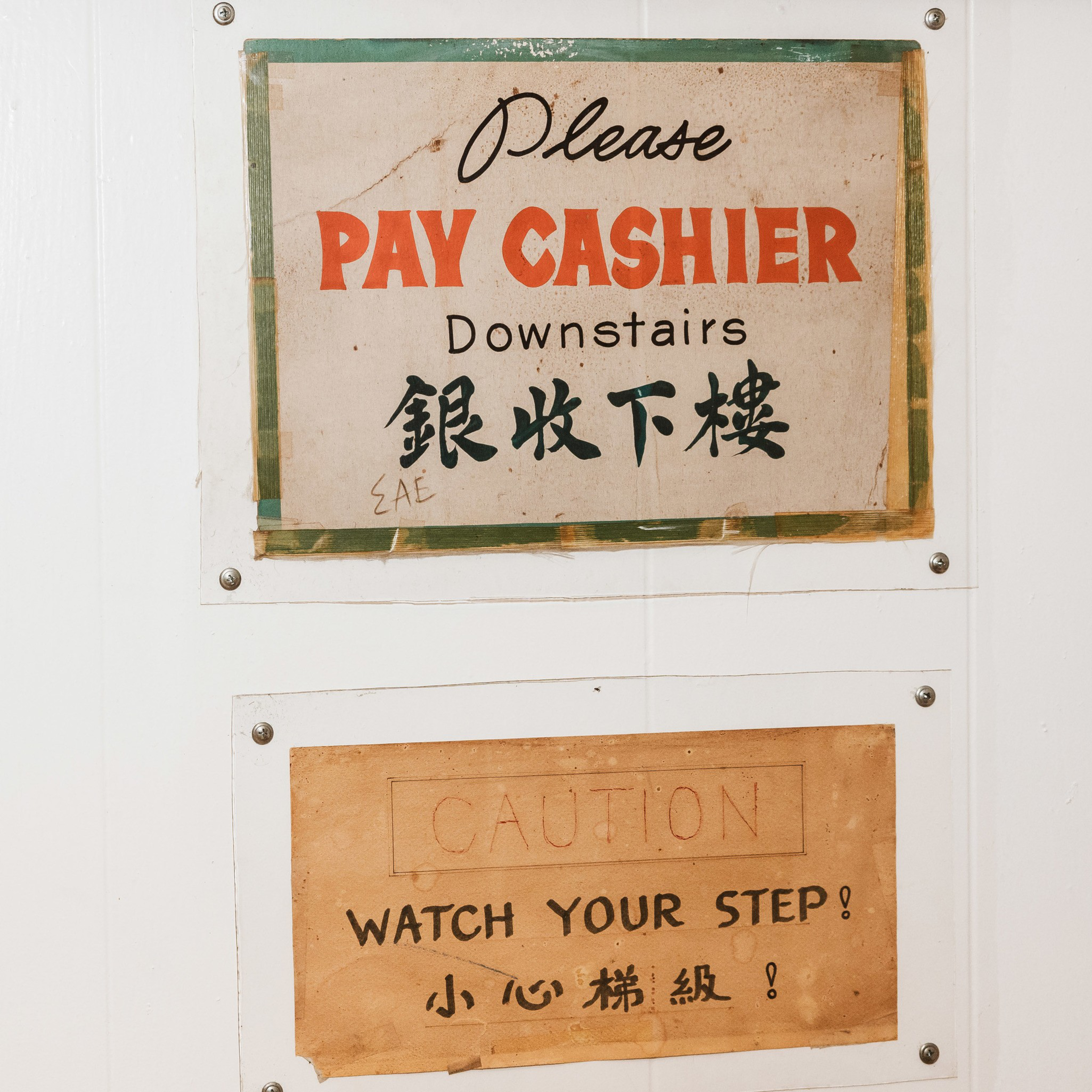 Two signs are posted on a wall. The top sign says, &quot;Please Pay Cashier Downstairs&quot; in English and Chinese. The bottom sign reads, &quot;Caution: Watch Your Step&quot; in both languages.