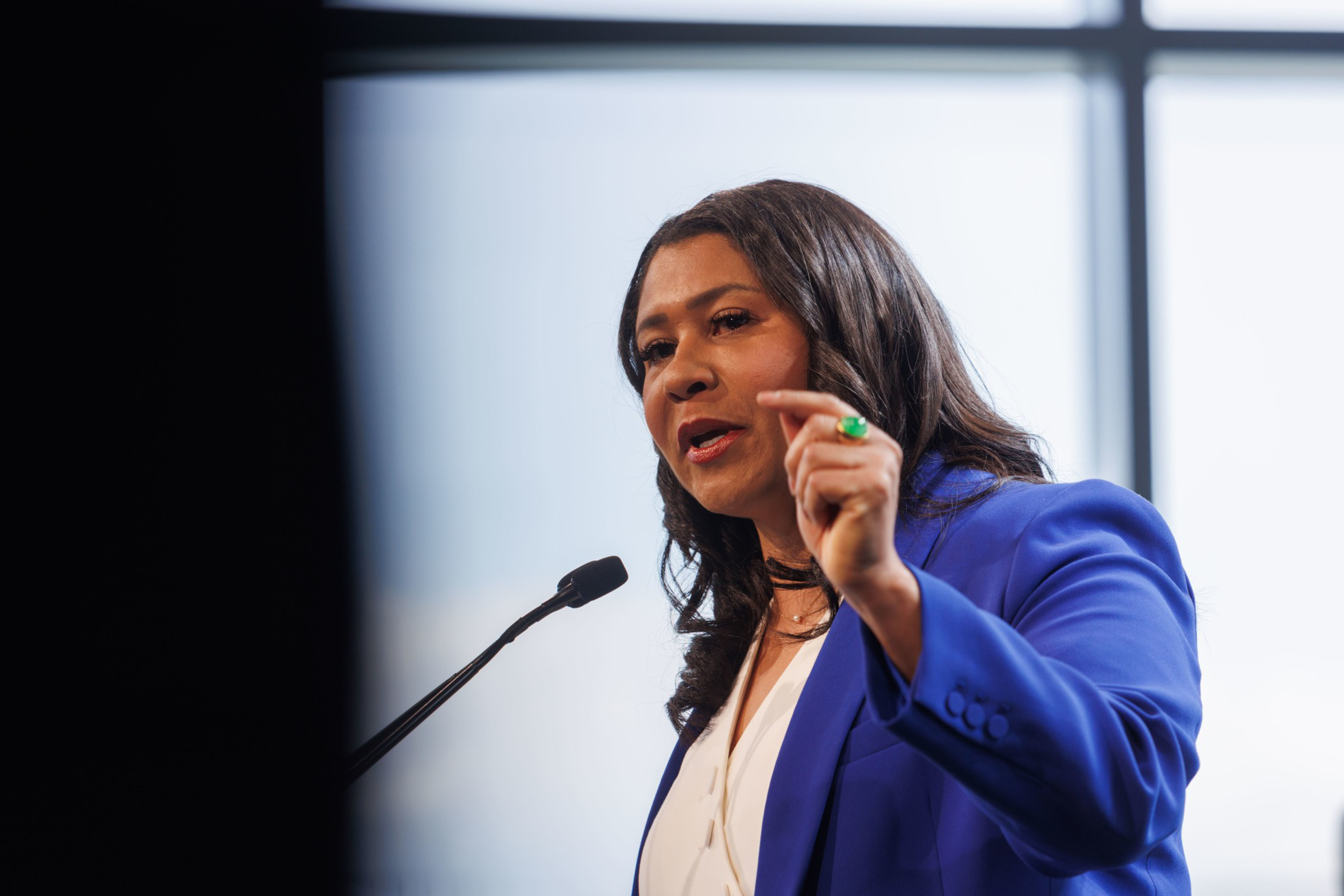A woman in a blue blazer gestures while speaking at a podium.