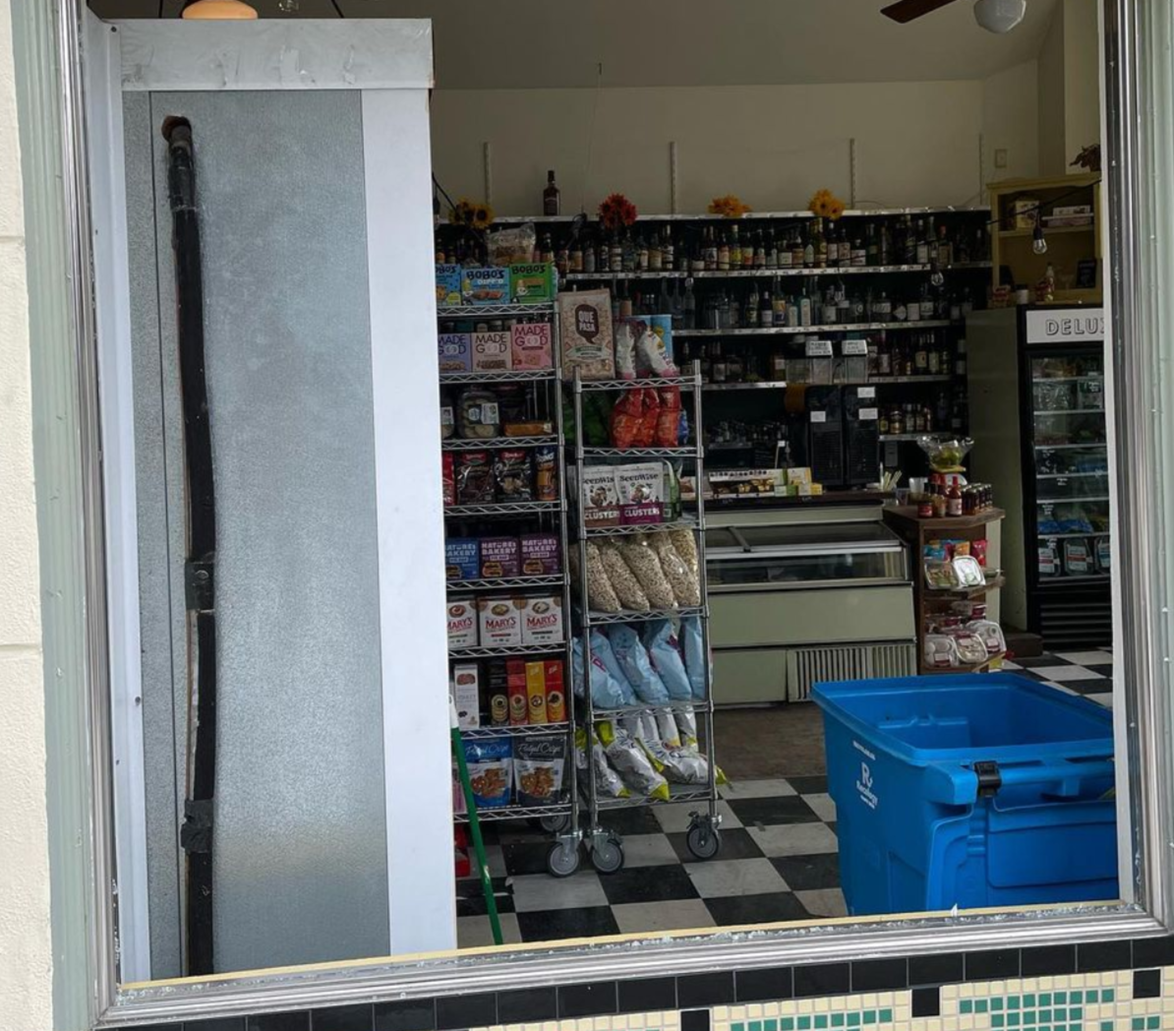 Shelves of snacks and drinks are visible inside a small store with a black and white checkered floor and a large blue bin in the foreground.