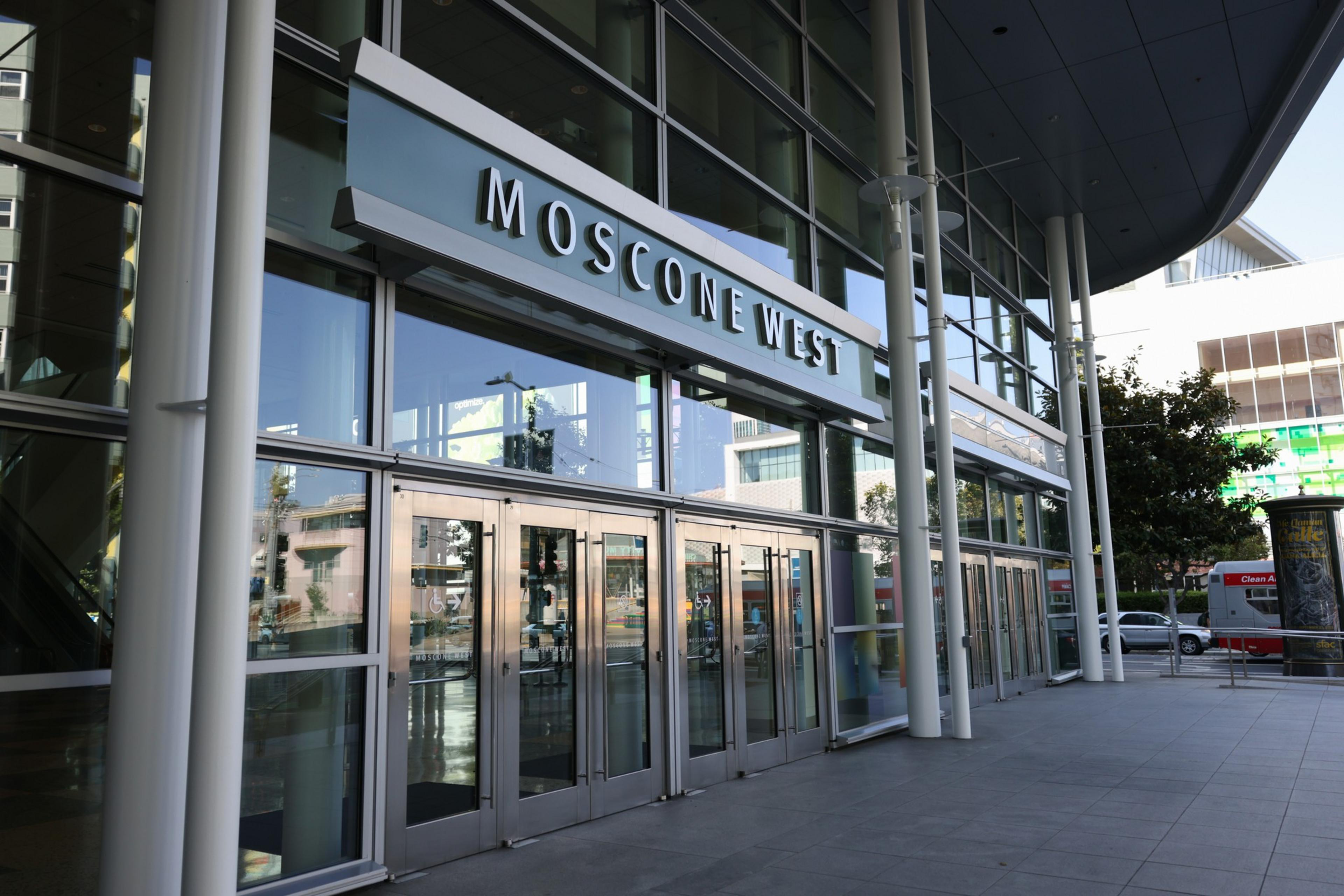 A modern glass building entrance with multiple glass doors beneath a sign reading "Moscone West," supported by tall white pillars along the sidewalk.