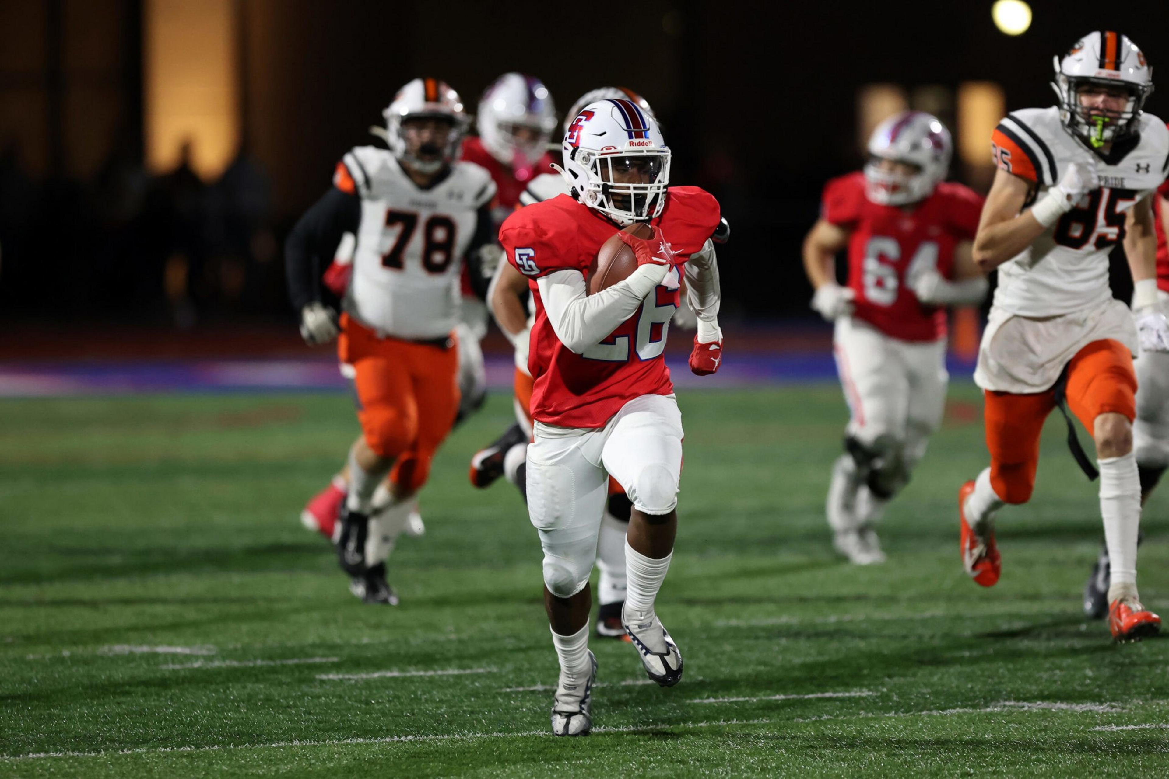 A football player in a red jersey and white pants runs with the ball while players in white and orange chase him on the field.