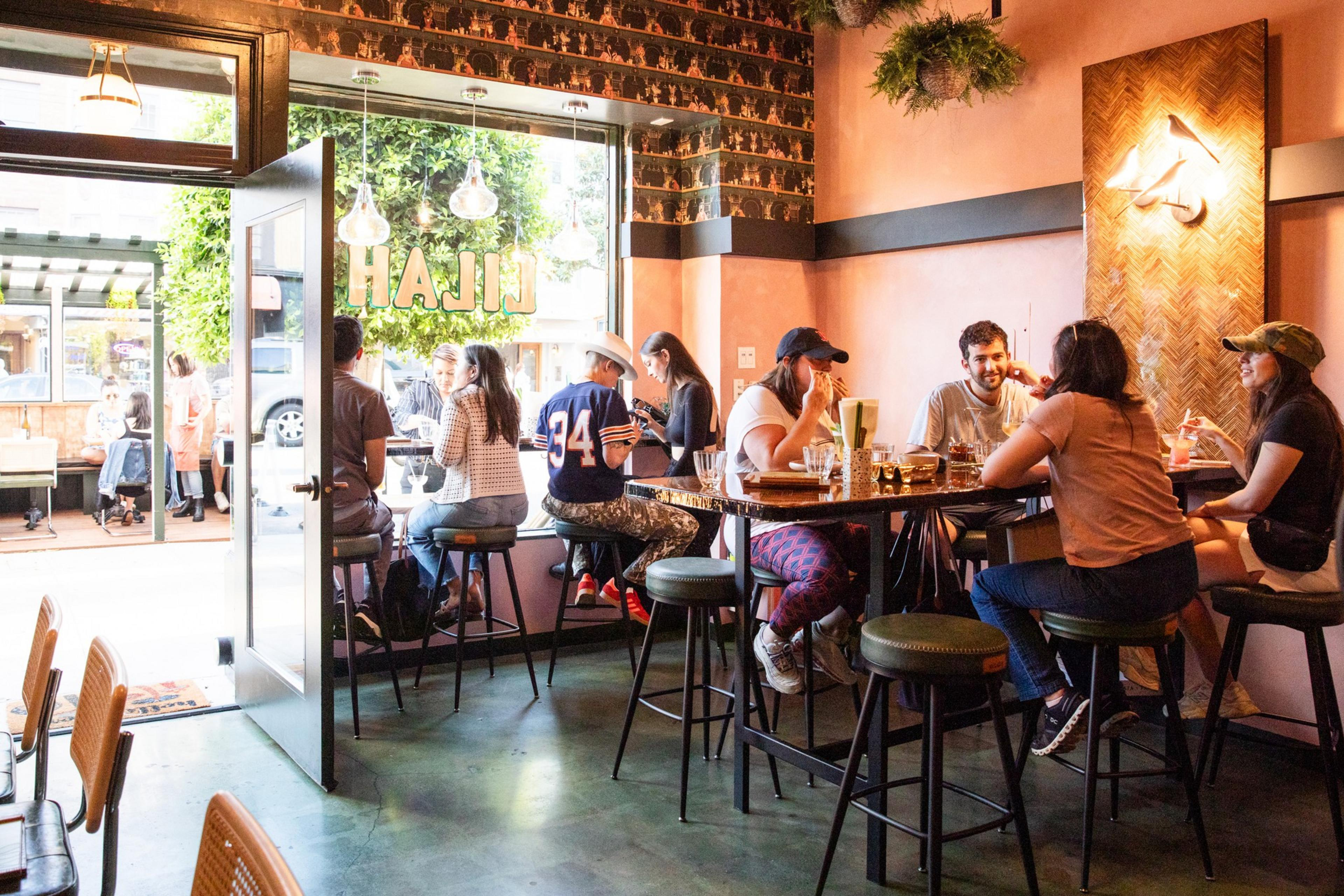 People are sitting at high tables inside a cozy cafÃ© with a warm ambiance. A few are near the open door enjoying drinks, and thereâ€™s greenery outside.