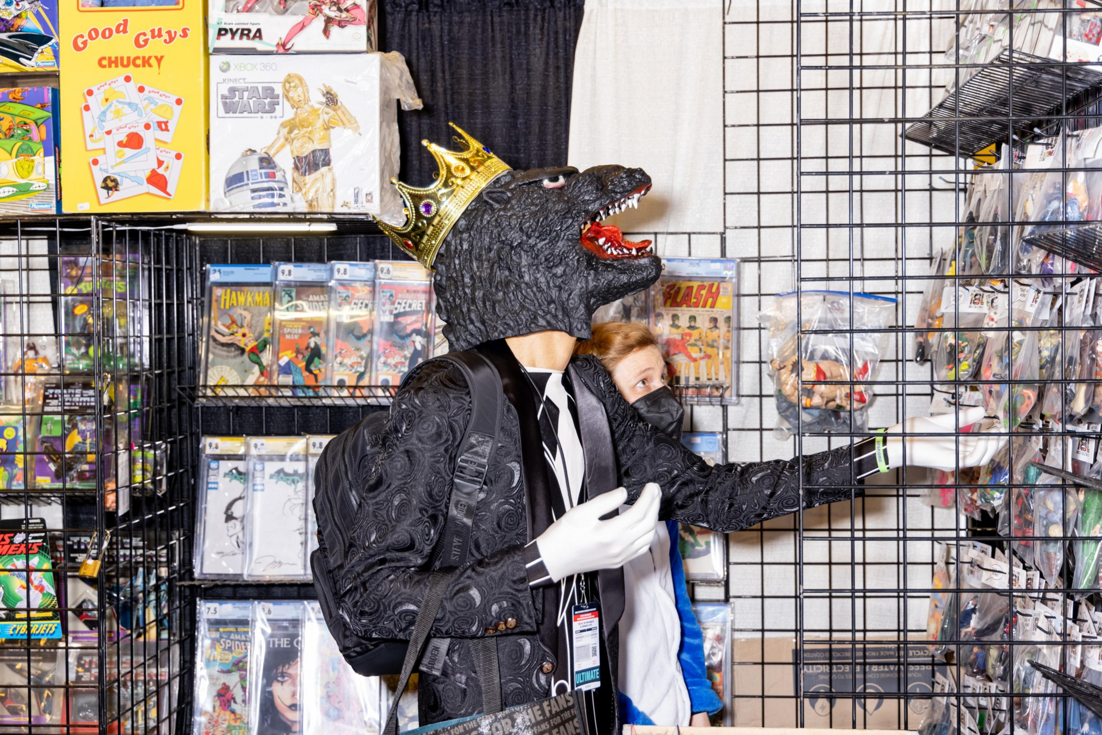 A person wearing a Godzilla mask and crown, dressed in a suit, reaches for items on a shelf at a comic book convention booth with comics and collectibles.