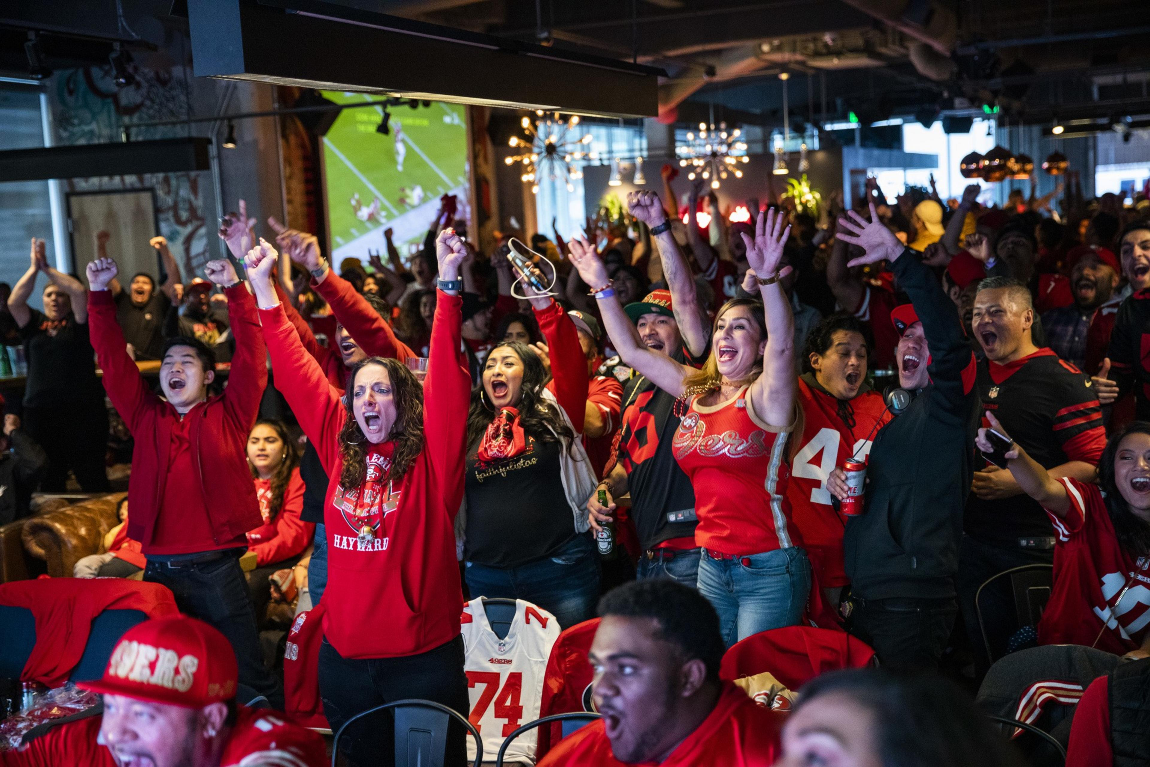A large group of people wearing San Francisco 49ers apparel jump and raise their hands in the air while celebrating inside a sports bar.