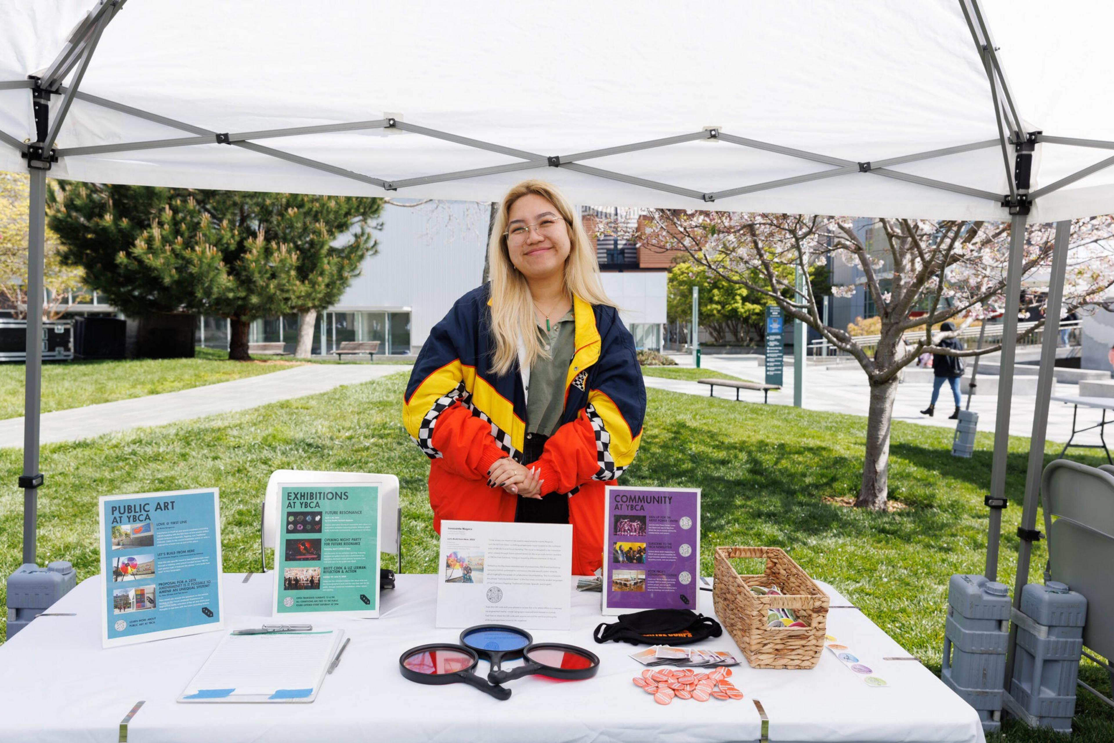A woman stands at an outdoor booth with pamphlets, a basket of goodies, and some pens.
