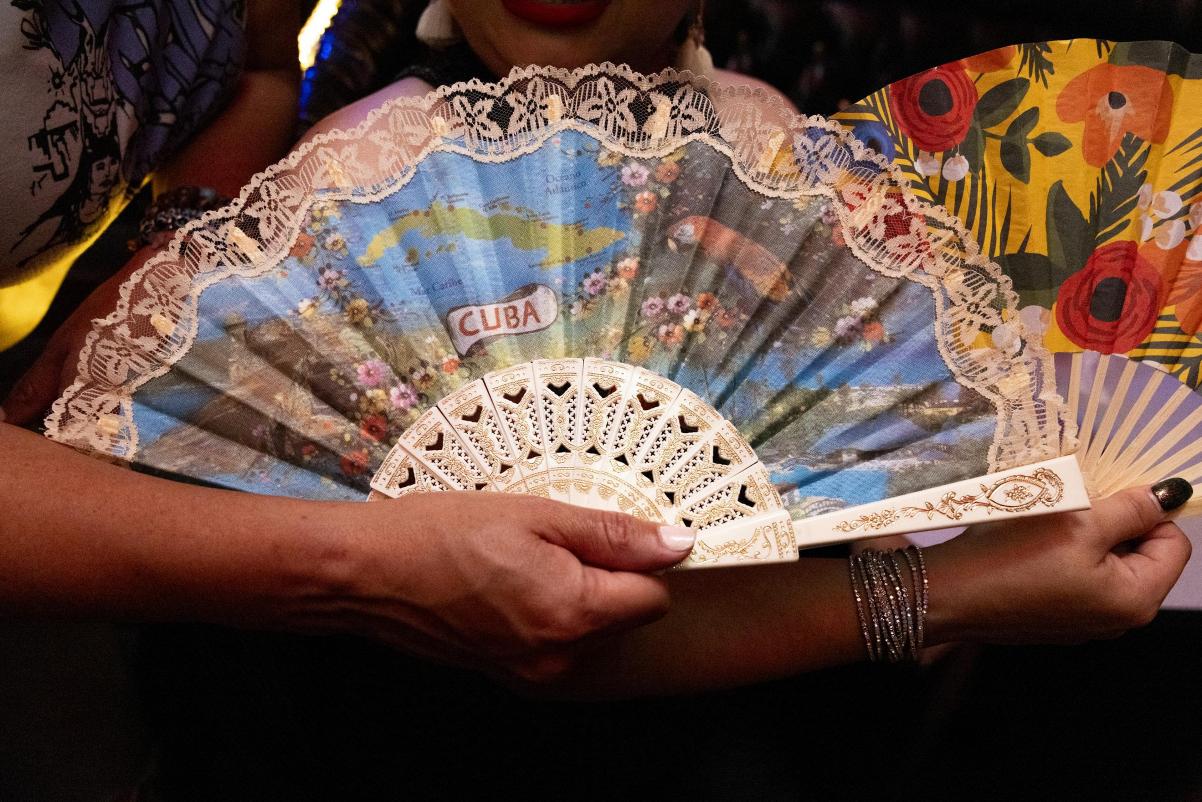 A person holds an intricately designed fan with a colorful map of Cuba and floral patterns, framed with lace. Behind it is another fan with a vibrant floral design.