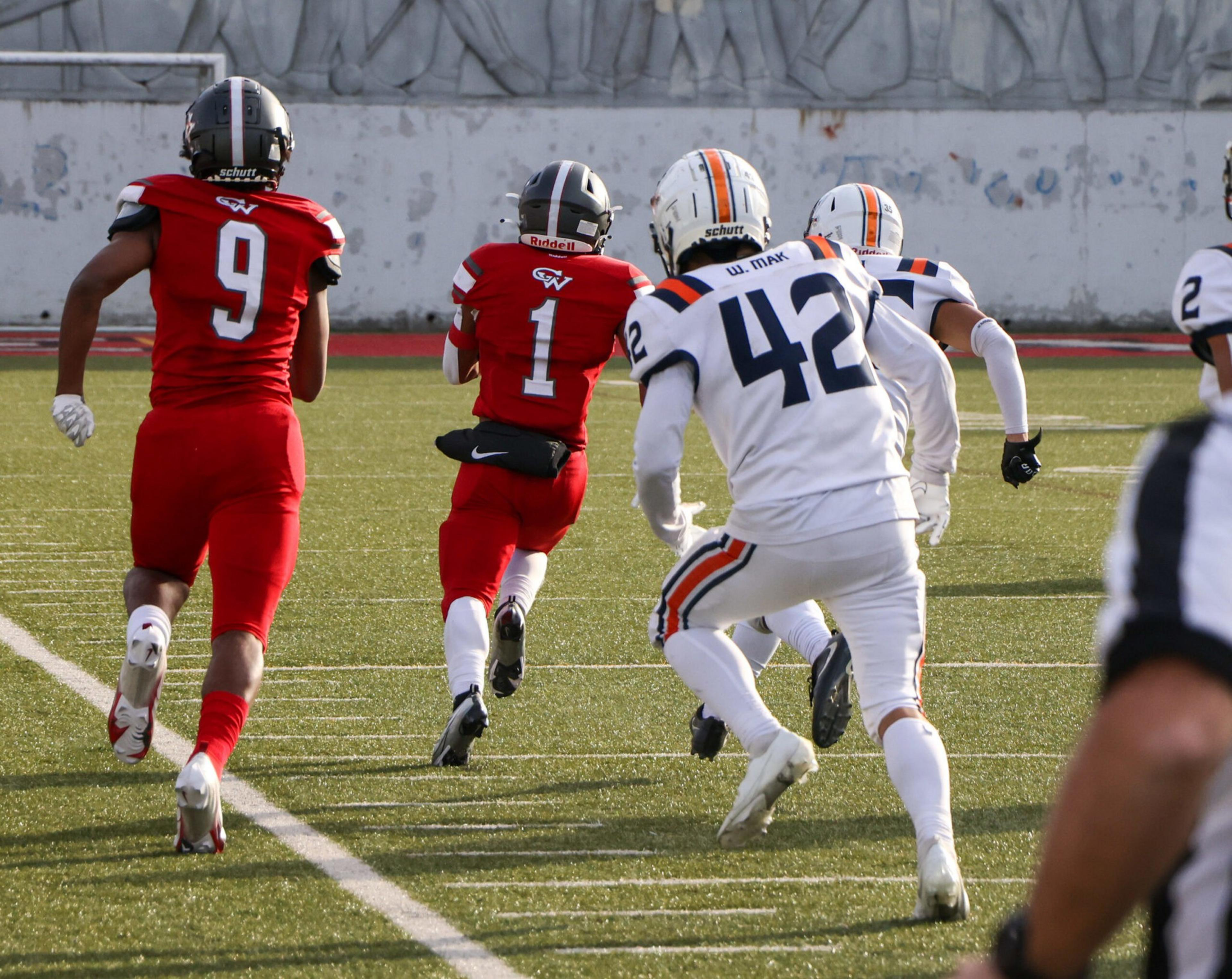 A football player in a red uniform runs with the ball while two players in white uniforms chase him on a green field.