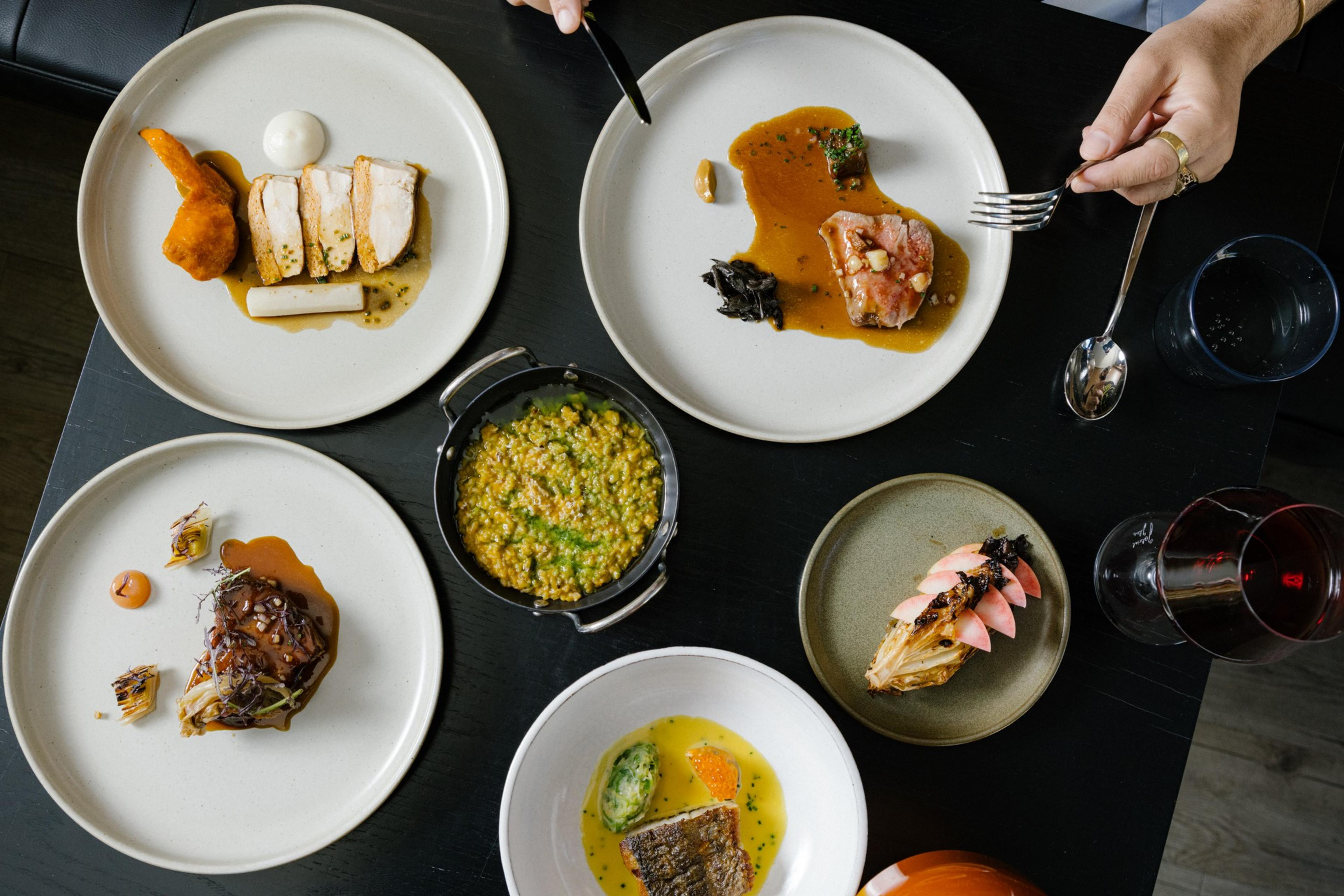 A variety of plated dishes with meats and vibrant sauces are arranged on a table, accompanied by a small pot of grain dish and a glass of red wine.