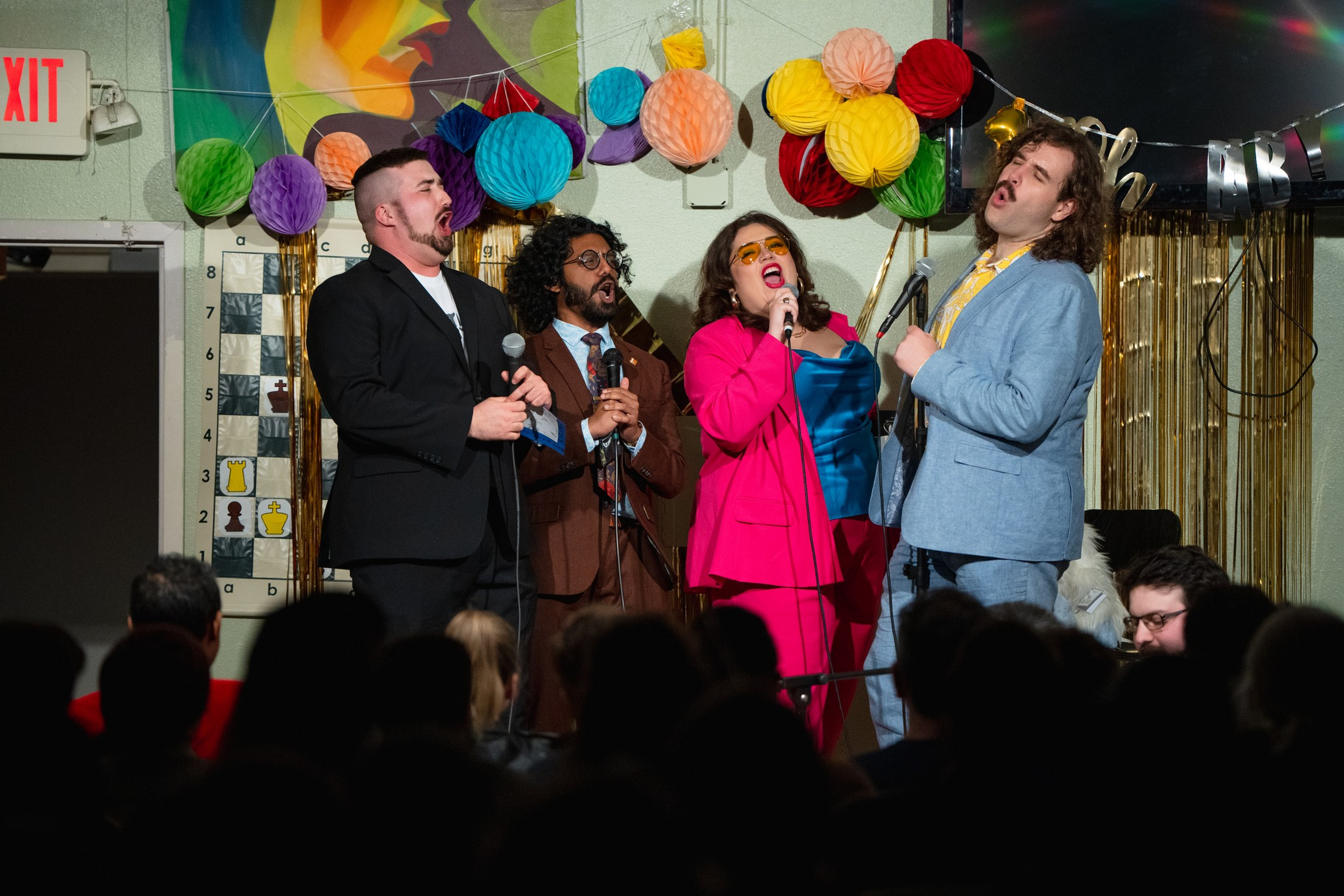 Four people dressed in colorful suits passionately sing into microphones on a decorated stage with paper lanterns hanging above.
