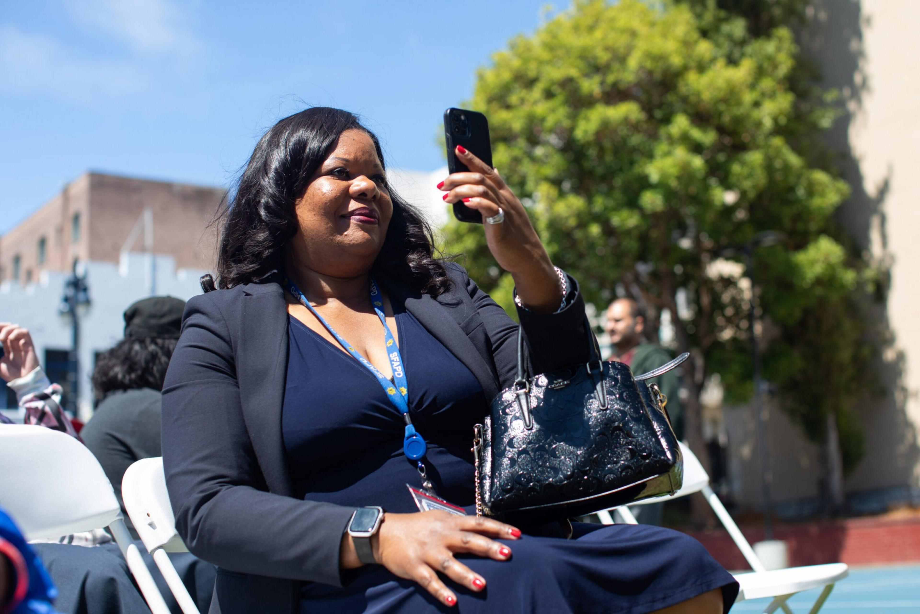 A woman in a navy dress and black blazer sits outdoors, holding a phone up for a selfie, with a black handbag on her lap and a lanyard around her neck.