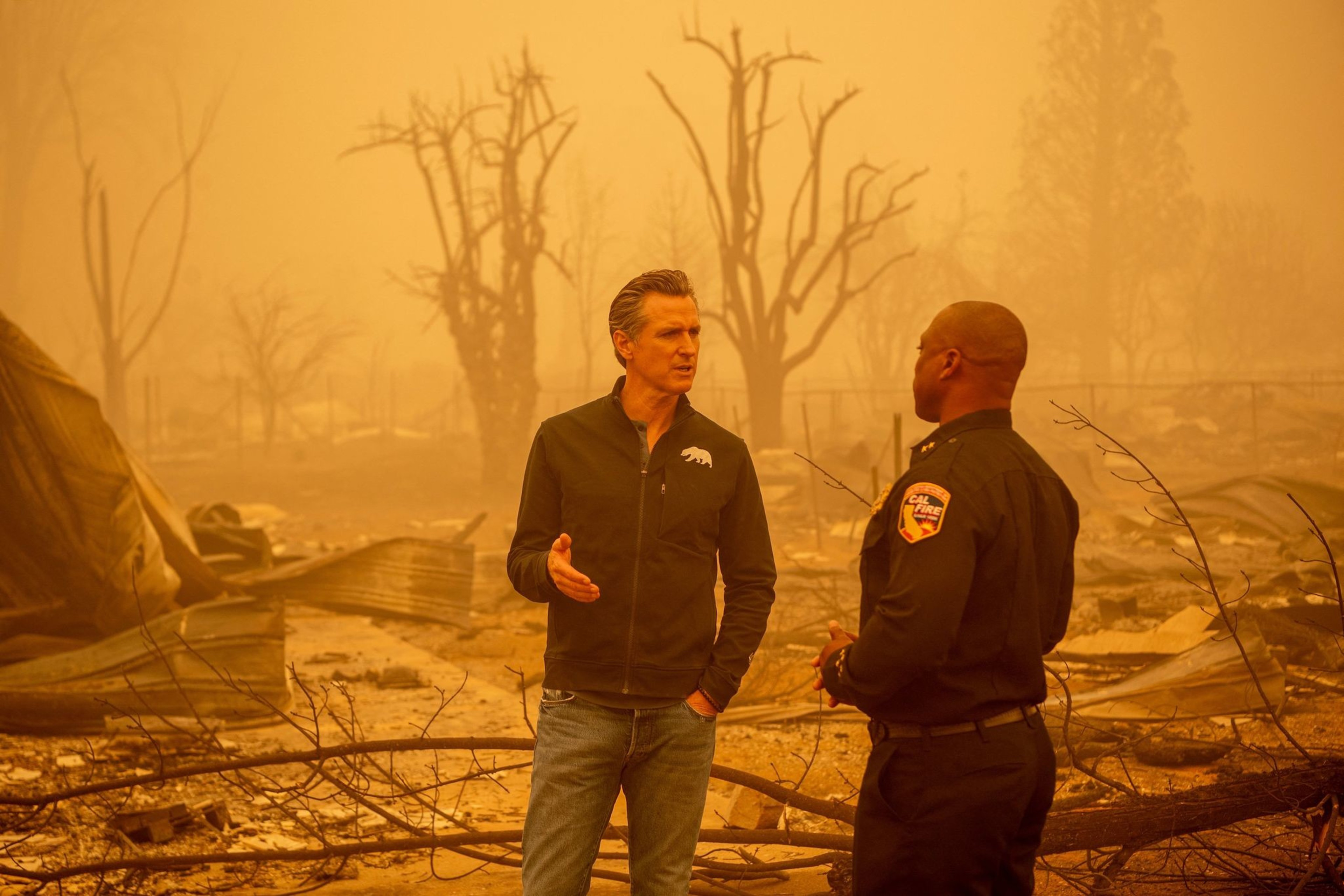 Two men stand talking in front a burnt landscape surrounded by orange haze.