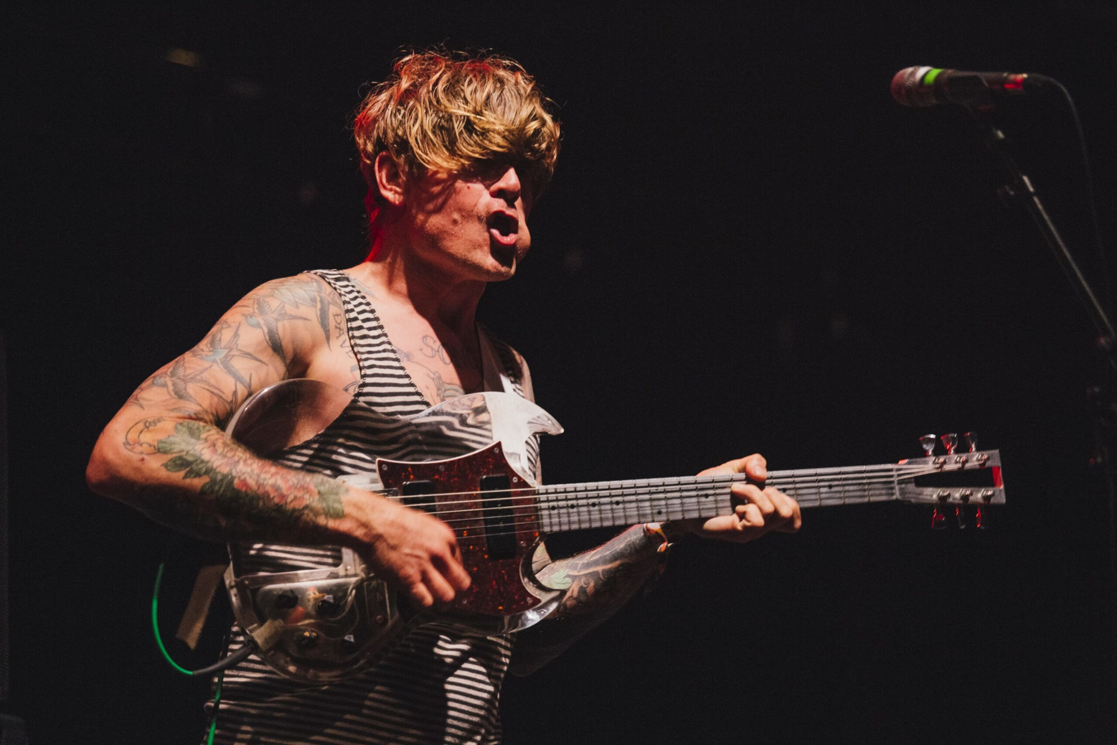A tattooed musician with tousled hair passionately plays an electric guitar and sings into a microphone on a dark stage.