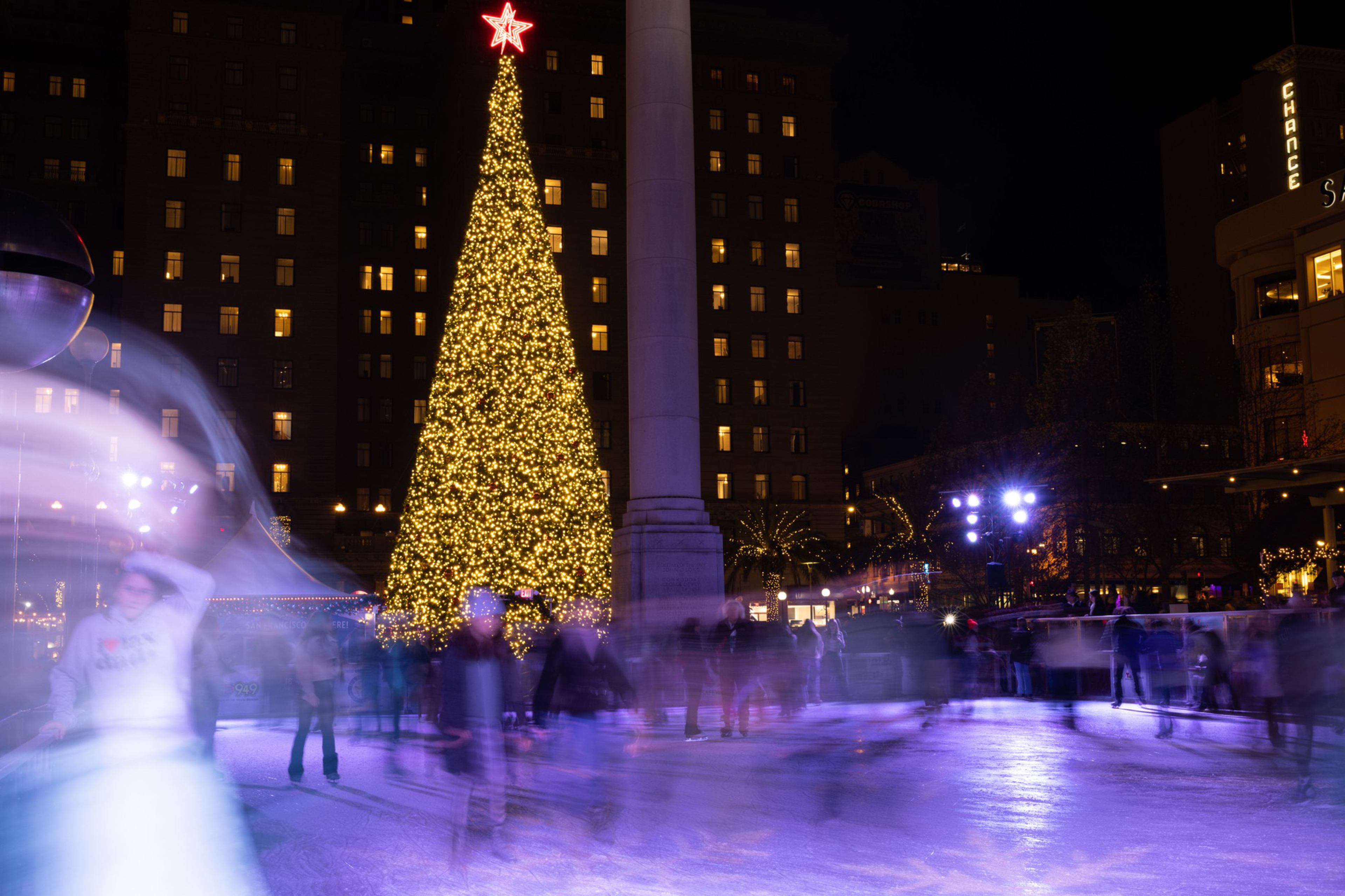 A large Christmas tree adorned with glowing lights stands prominently in a city square, surrounded by people ice skating under the night sky.