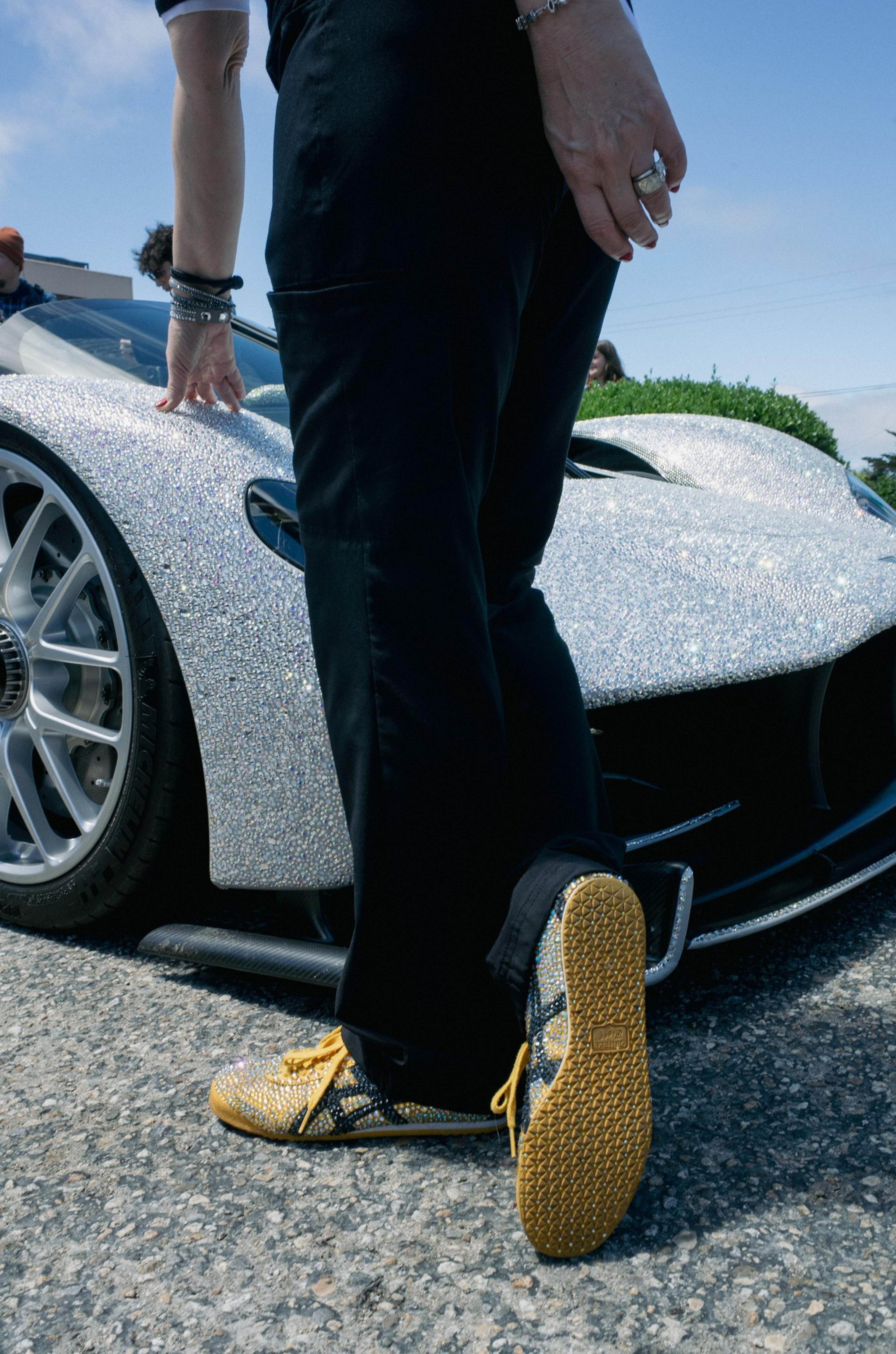 A person wearing shiny gold and black shoes touches a sparkling silver car with sleek, aerodynamic design and large wheels.