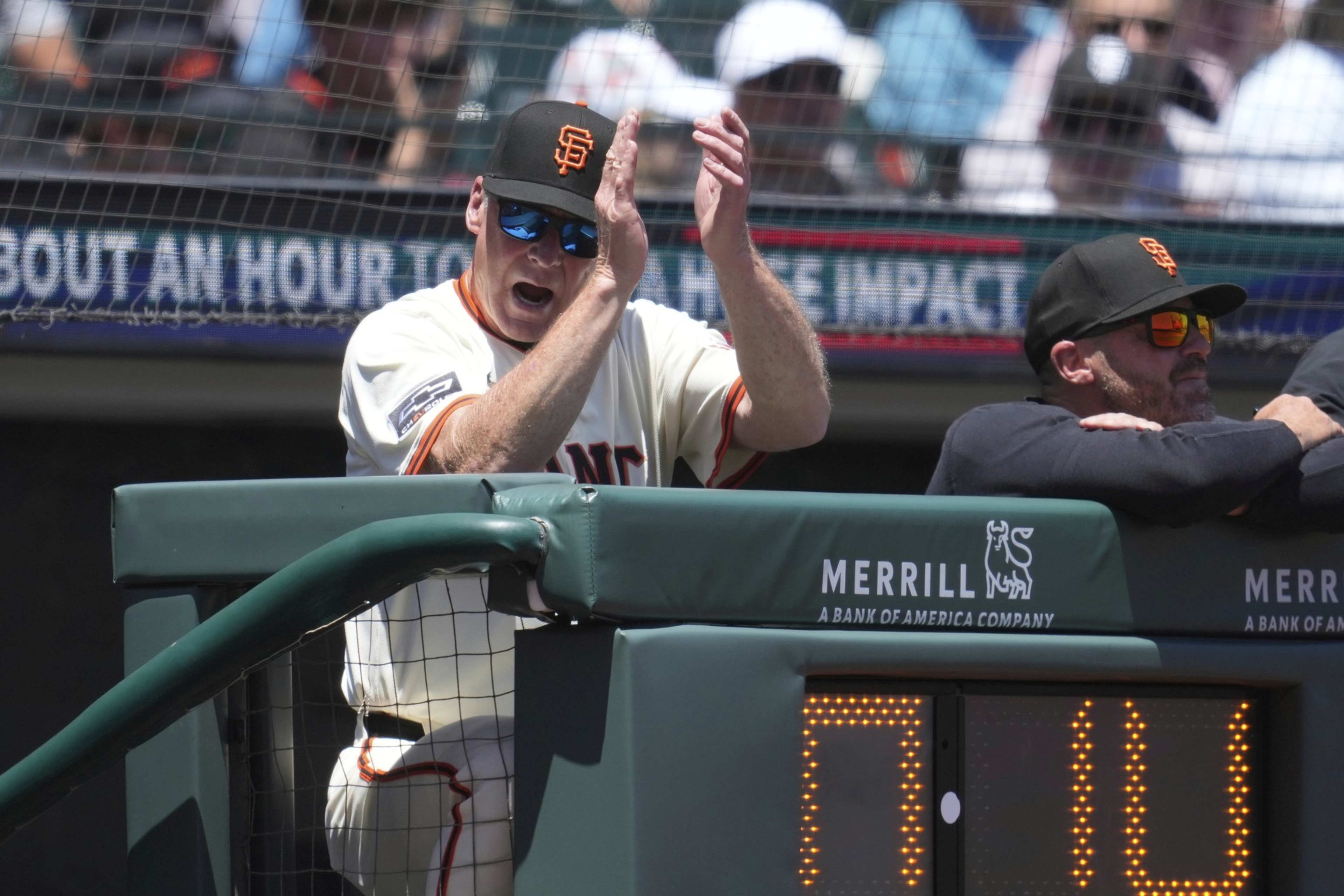 A baseball coach in a Giants uniform claps enthusiastically. He wears sunglasses and a cap, standing near the dugout railing with another person beside him.