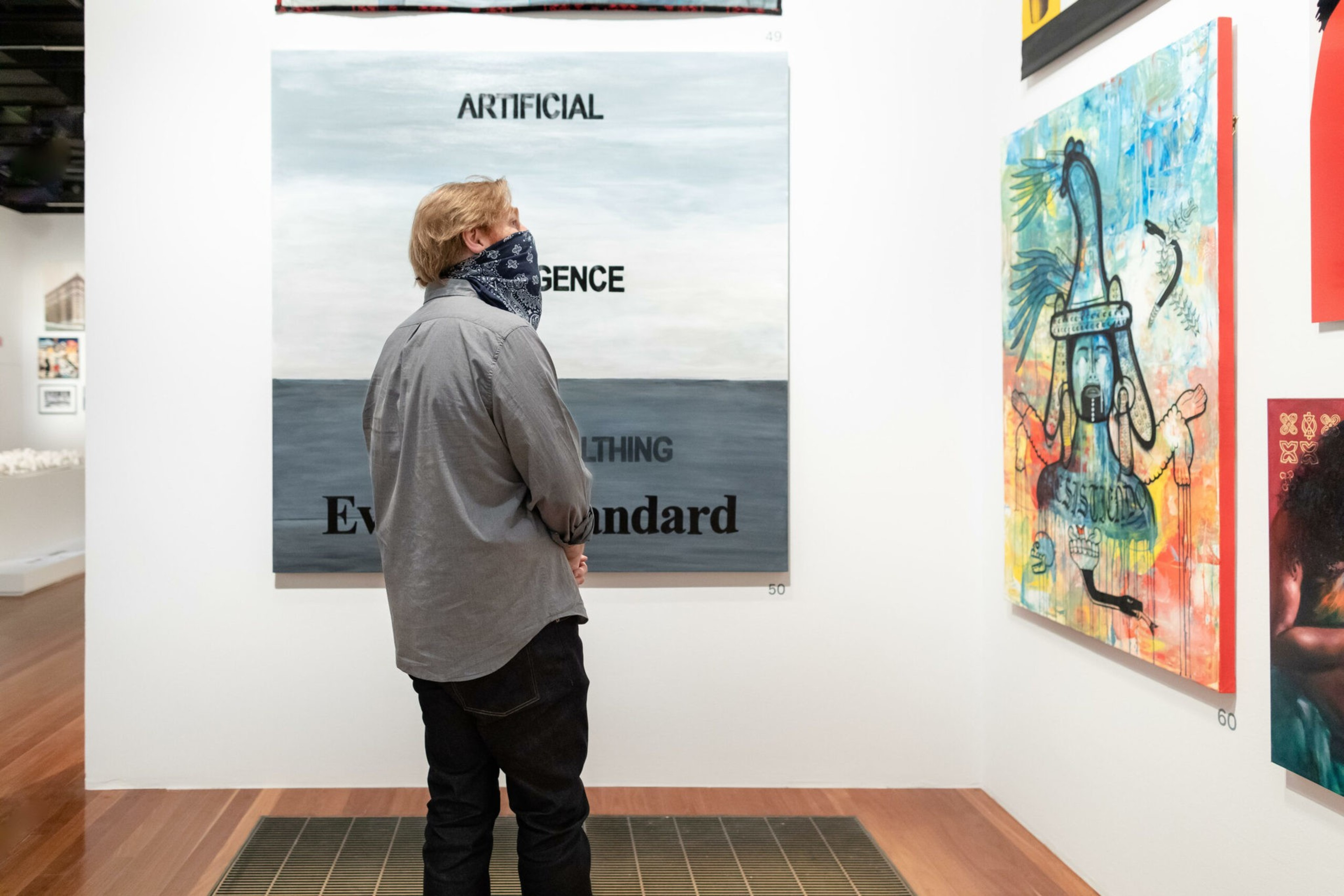 A person wearing a gray shirt and black pants, with a bandana covering their face, stands in an art gallery observing a large painting with text.