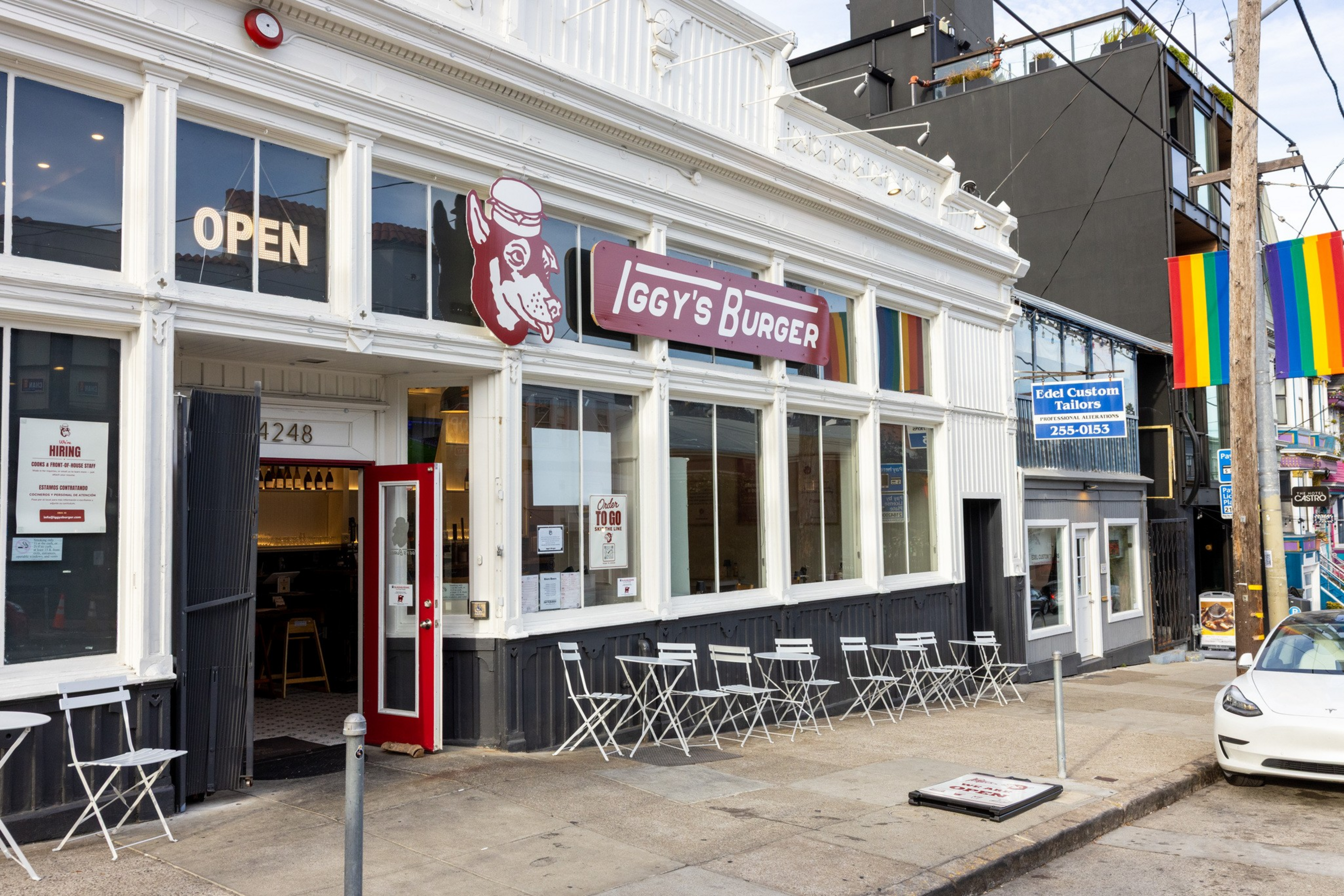 Iggy's Burger storefront features white and black exterior walls, red door, outdoor seating with white chairs and tables, and two rainbow flags on the street.