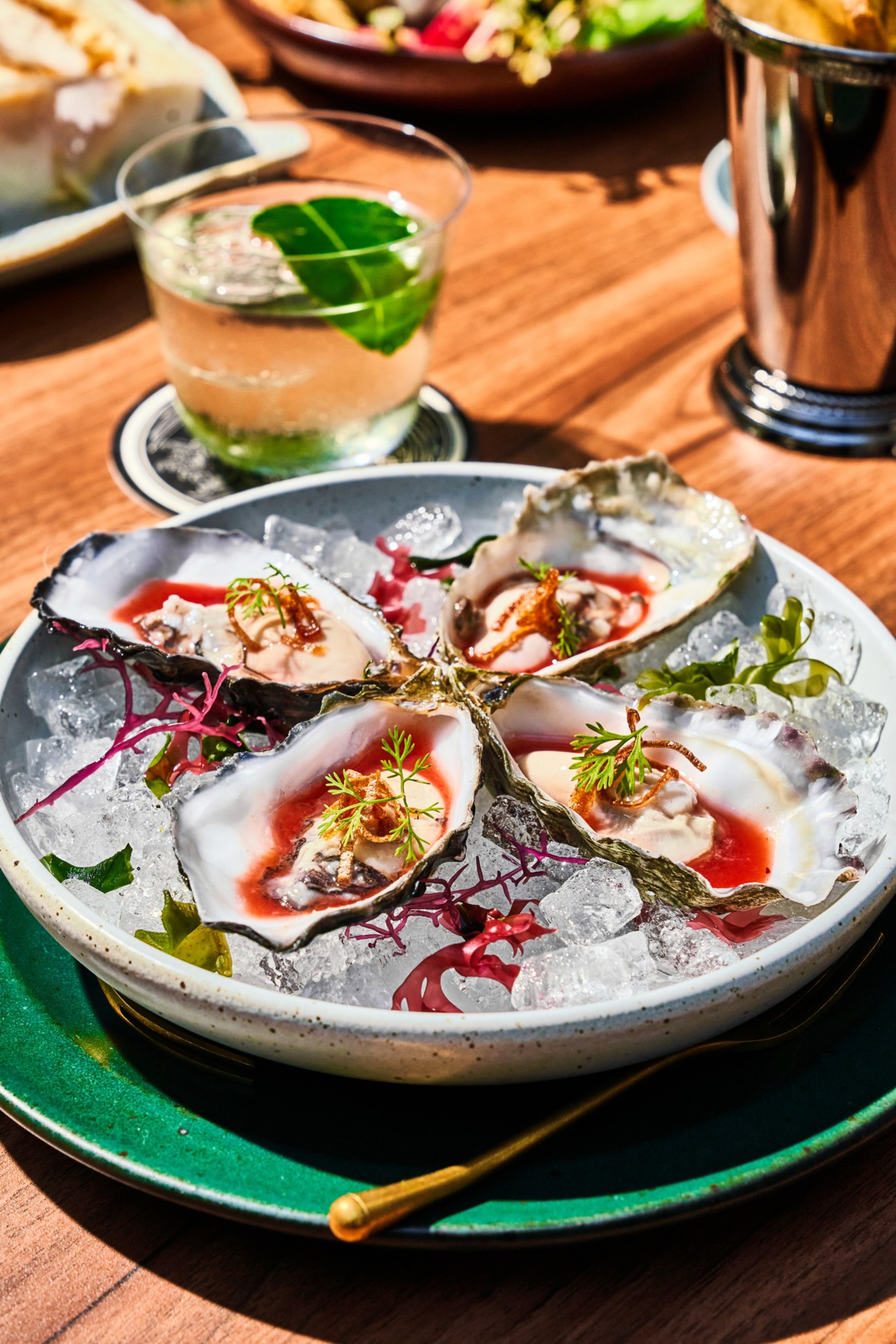 A plate of oysters on ice is garnished with herbs and accompanied by a cocktail with a leaf floating inside, set on a wooden table.