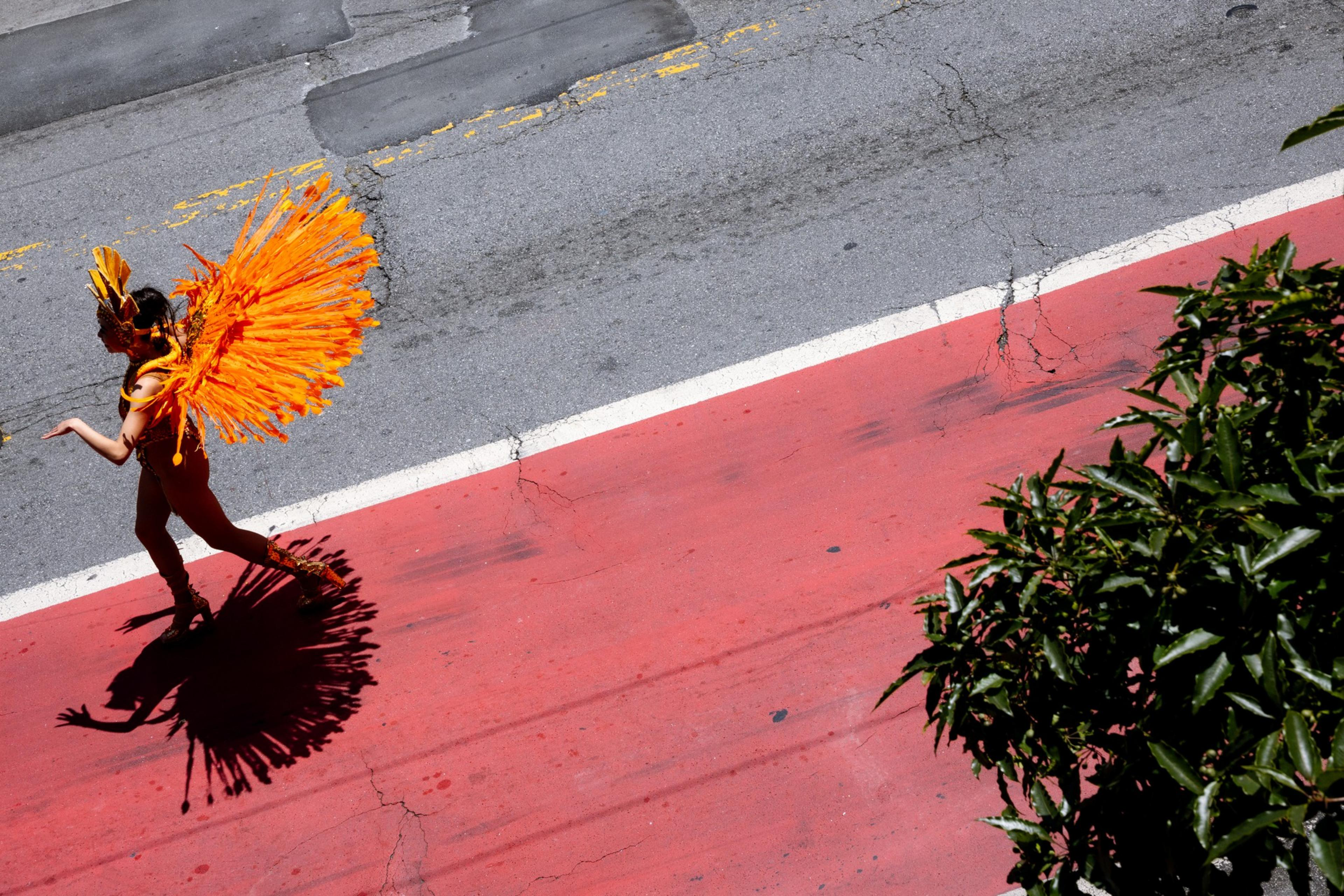 A person in vibrant orange feathered attire and a matching headdress walks on a red road. Their shadow is prominent, and greenery is seen on the right.