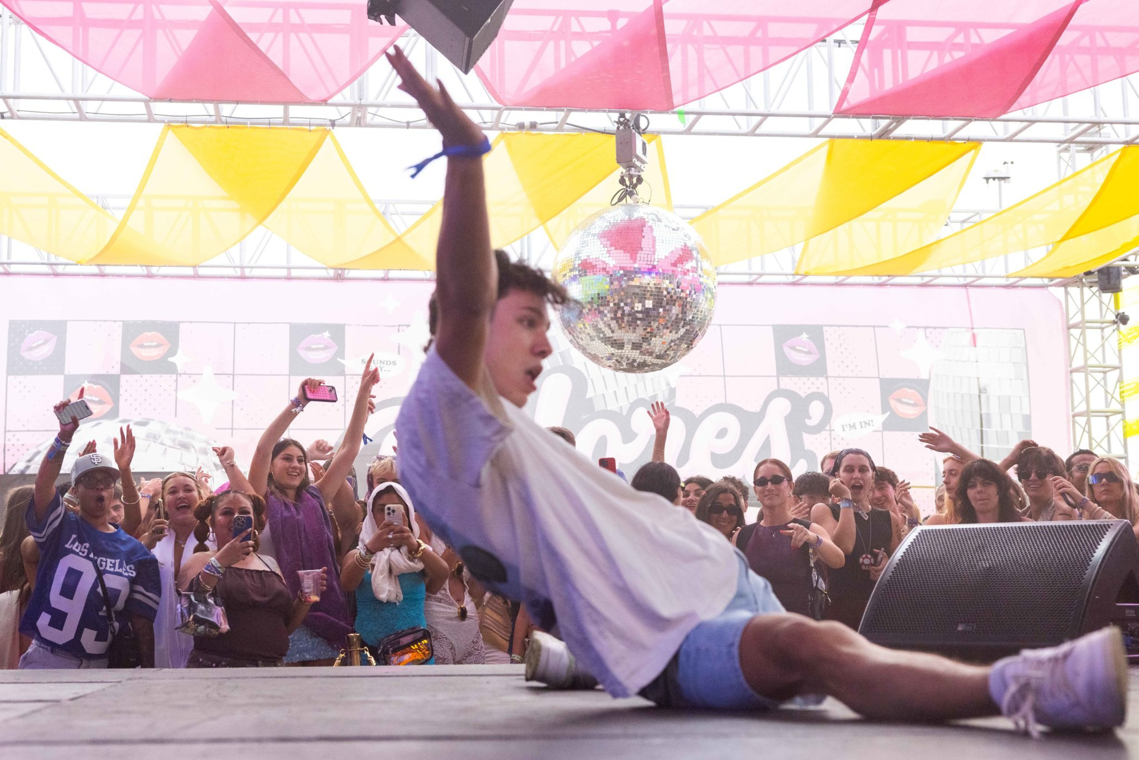 A young man slides on stage with arms raised while a crowd behind him cheers and takes photos under pink and yellow fabric decorations.