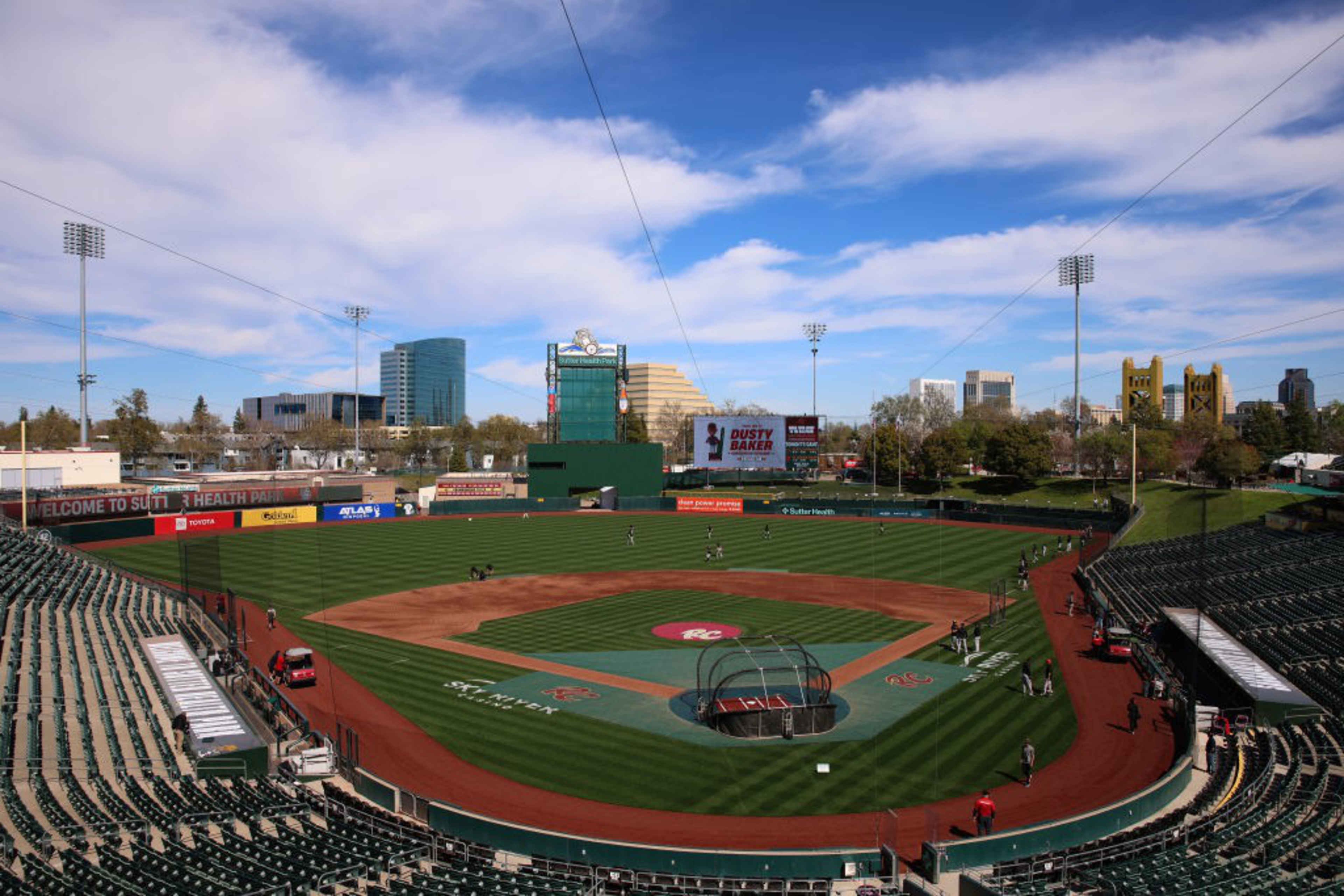 A baseball stadium with an empty field and neatly mowed grass is seen under a bright blue sky. Bleachers surround the field, with a city skyline in the background.