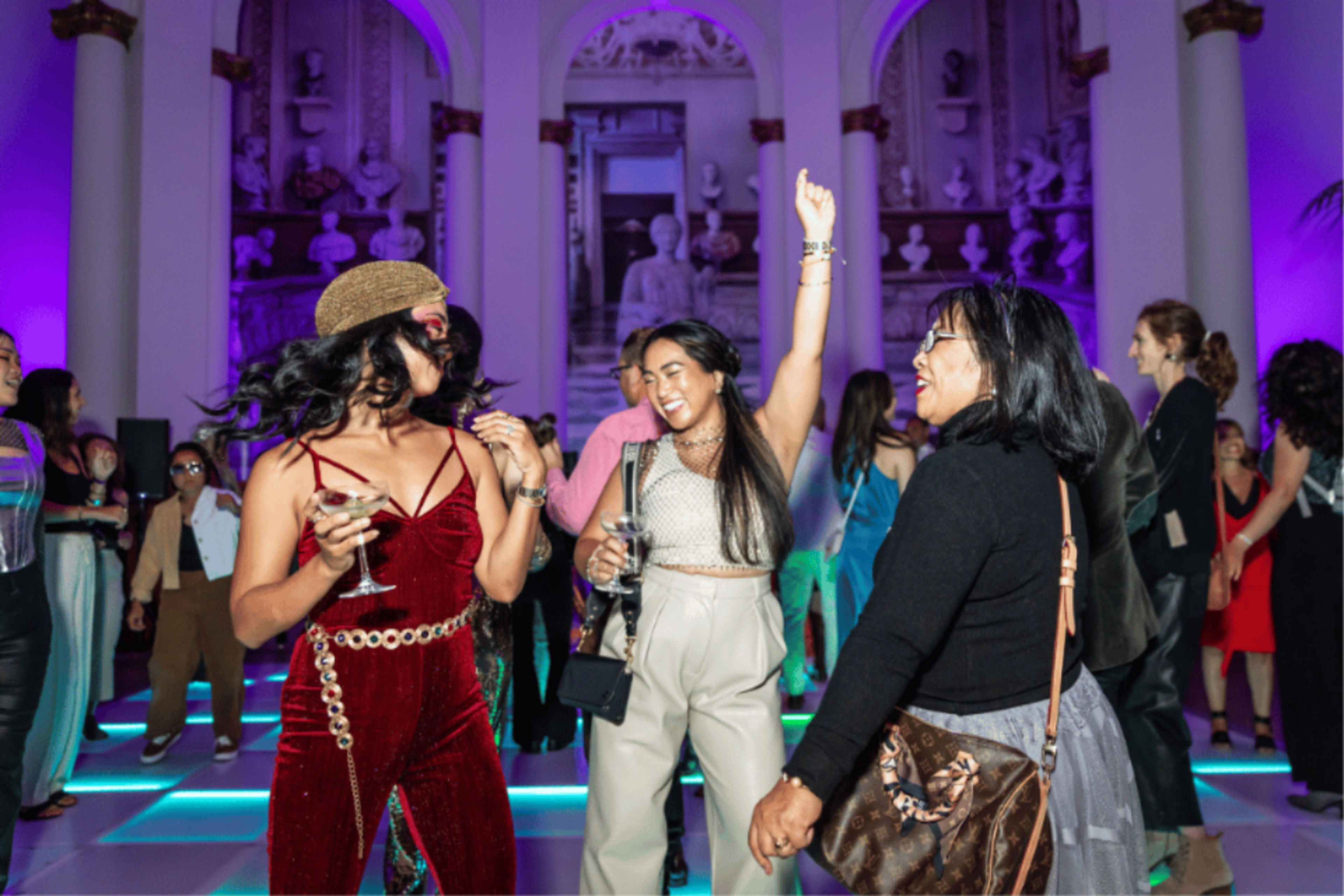 Three women are joyfully dancing in a vibrant room with purple lighting, surrounded by other people. One holds a drink, while another has her arm raised.