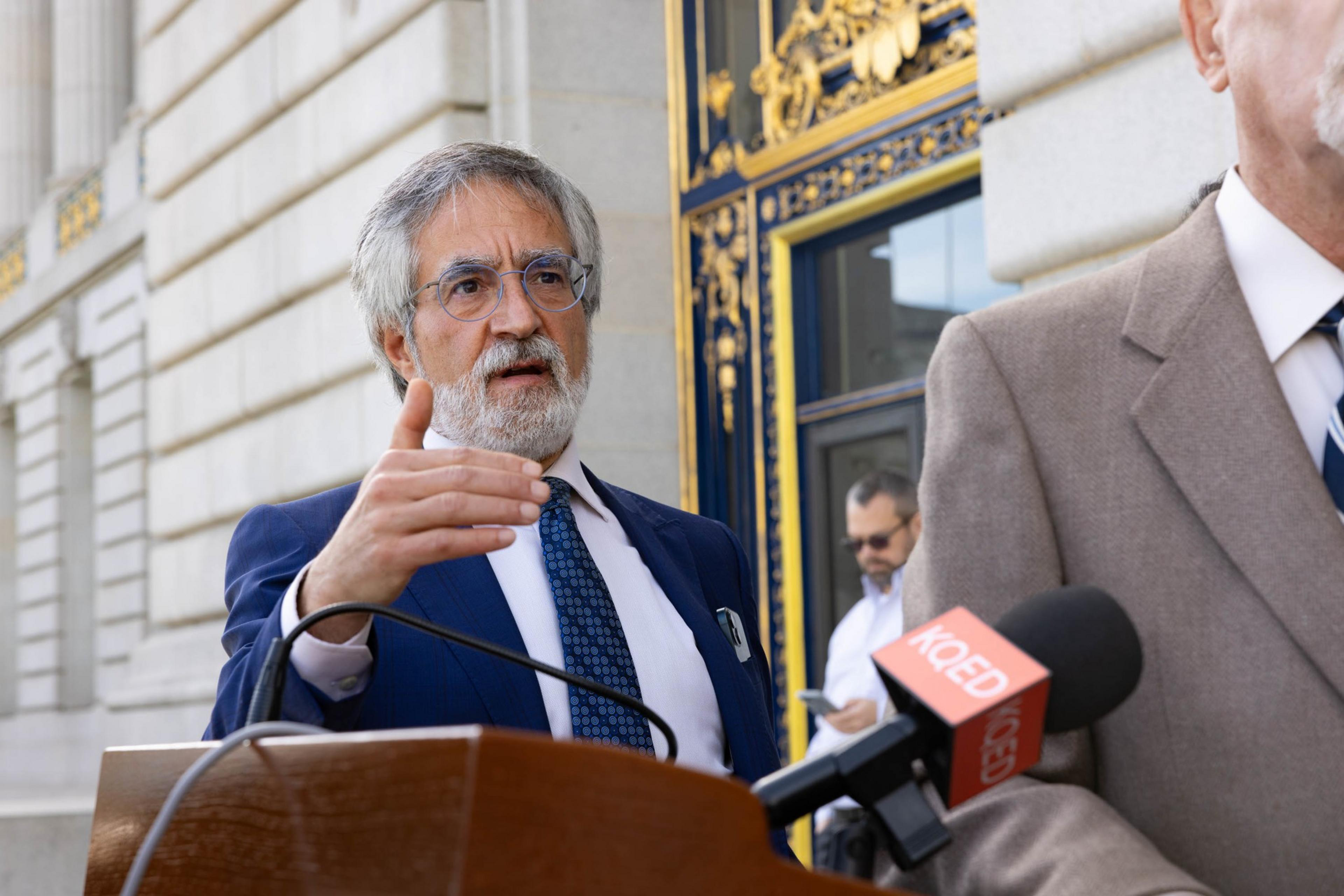 A man gesticulates and speaks with 1 hand extended out next to a podium with a gilded facade in the background.