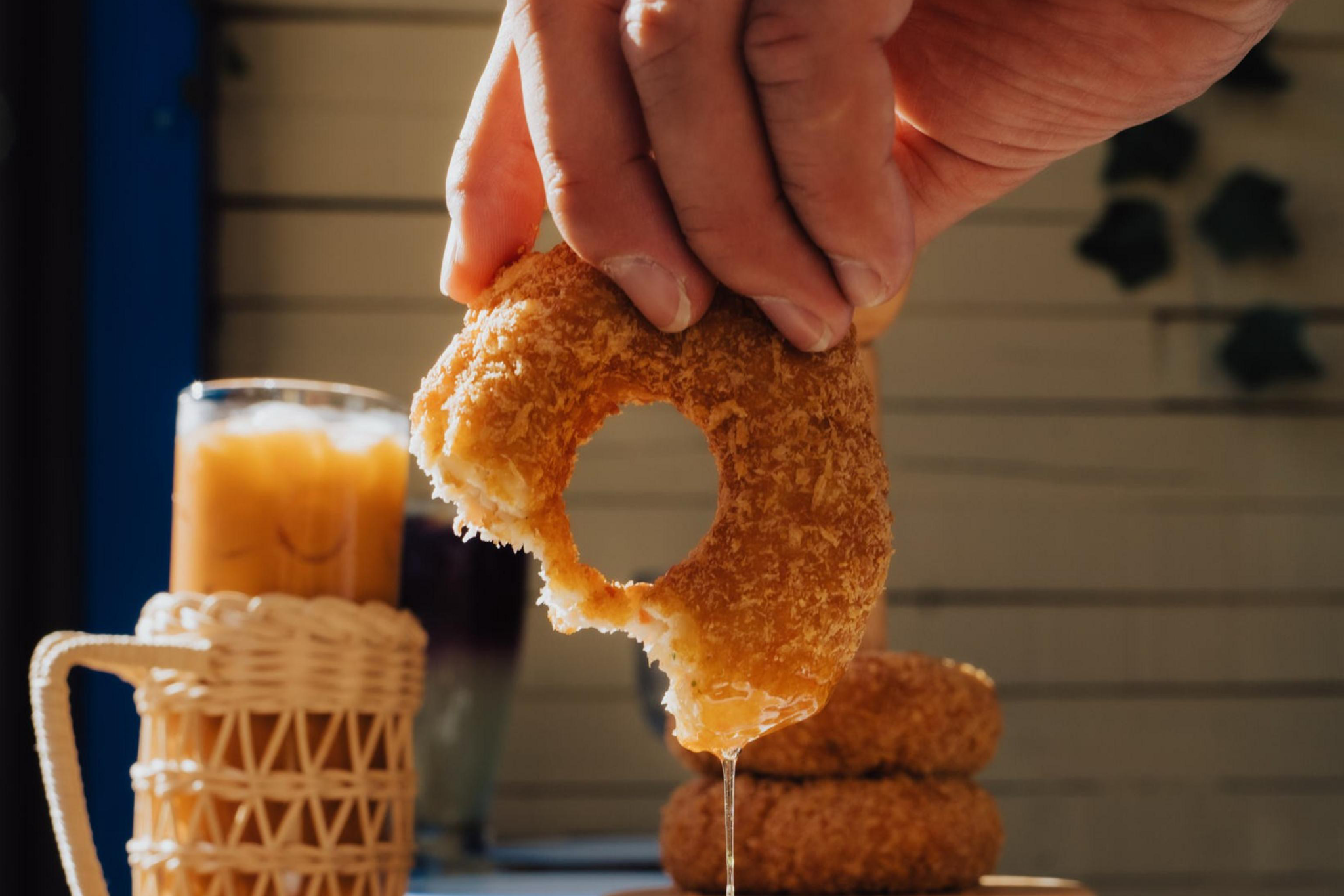 A hand holds a bitten, round fried donut dripping sauce over a small white bowl on a plate, with iced drinks and more donuts in the background.