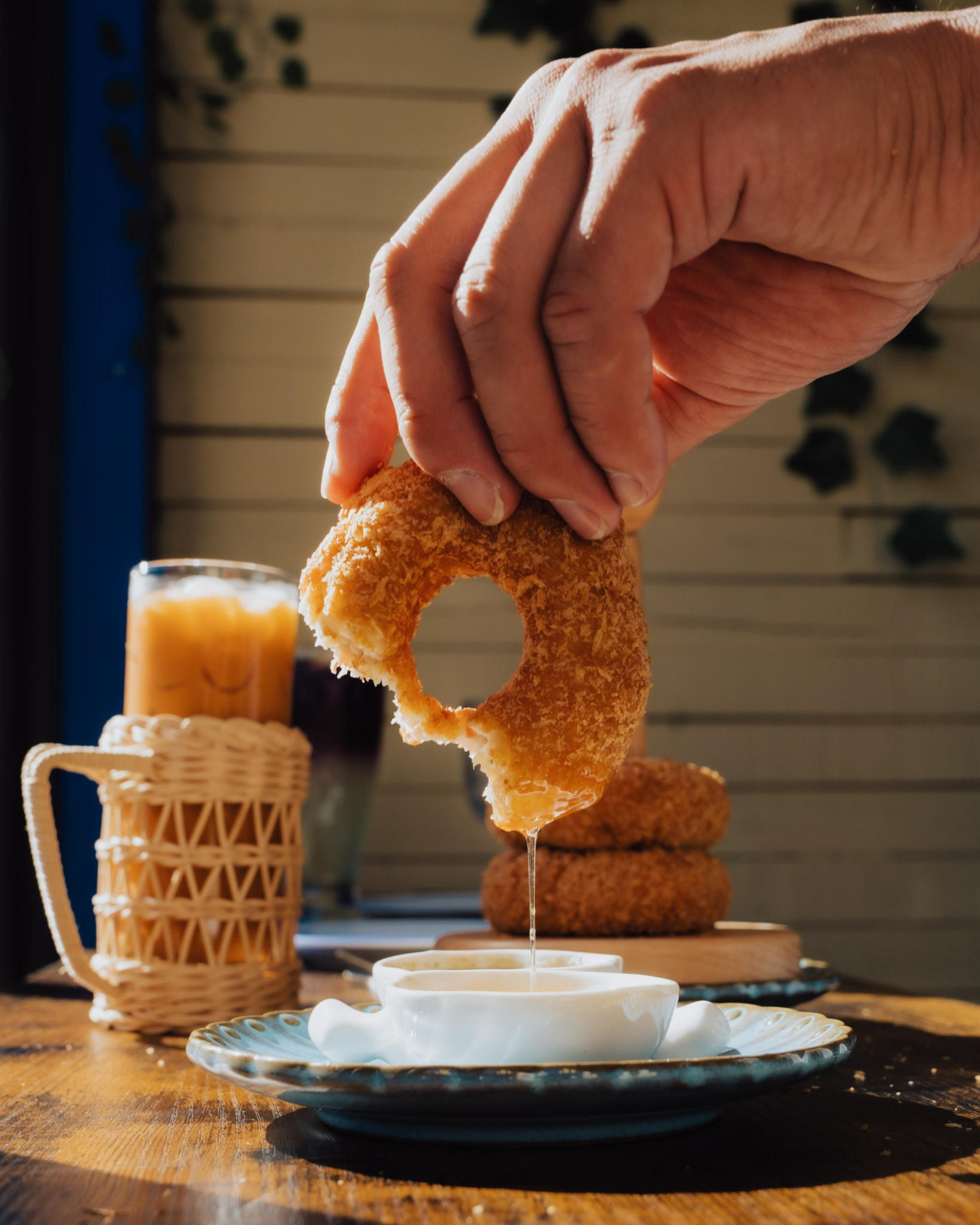 A hand holds a bitten, round fried donut dripping sauce over a small white bowl on a plate, with iced drinks and more donuts in the background.