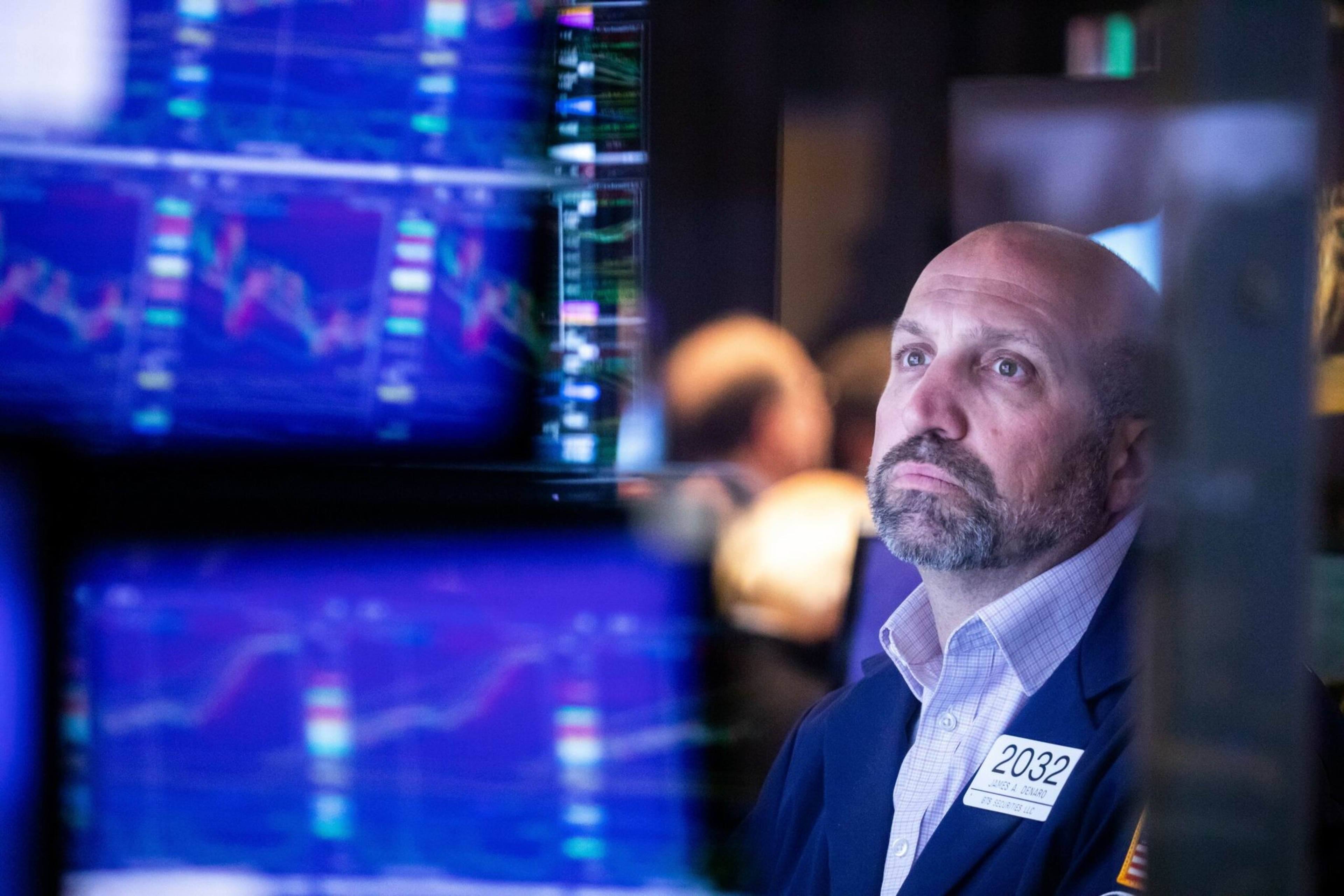 A bald man with a beard in a blue jacket stares thoughtfully at multiple blurred computer screens displaying colorful stock market graphs.