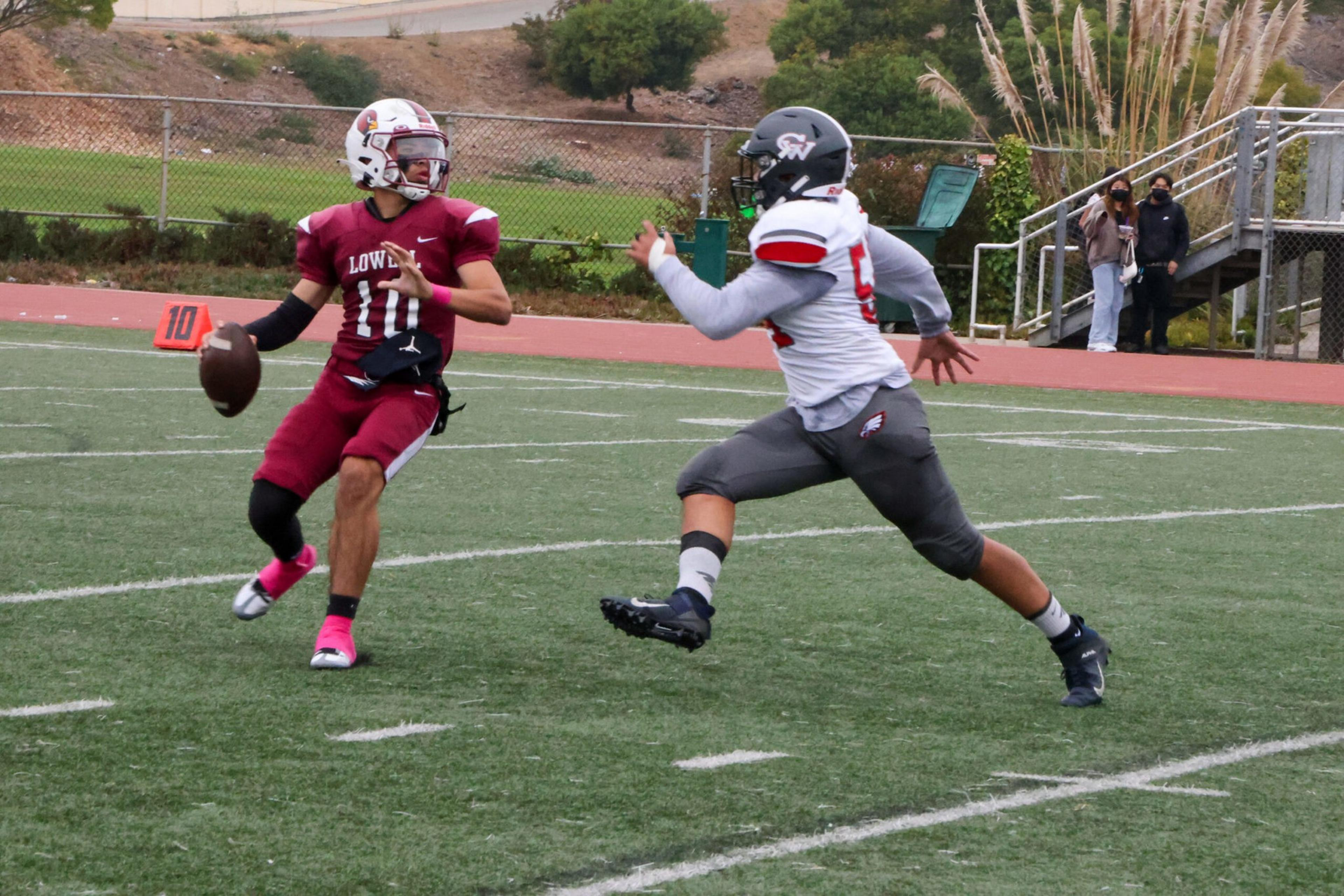 A football player in maroon prepares to throw the ball while a player in white and gray rushes to tackle him on a green field.
