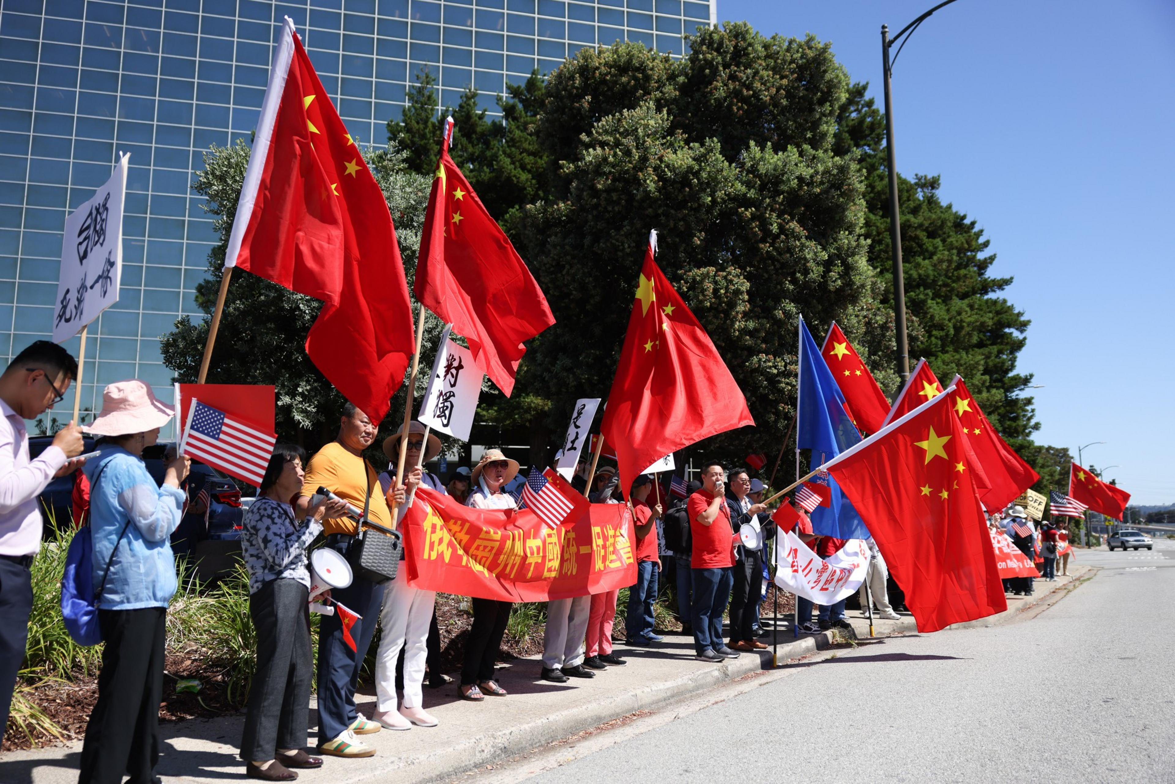 People stand on a sidewalk holding Chinese and American flags, banners, and signs, with a large building and trees in the background on a sunny day.