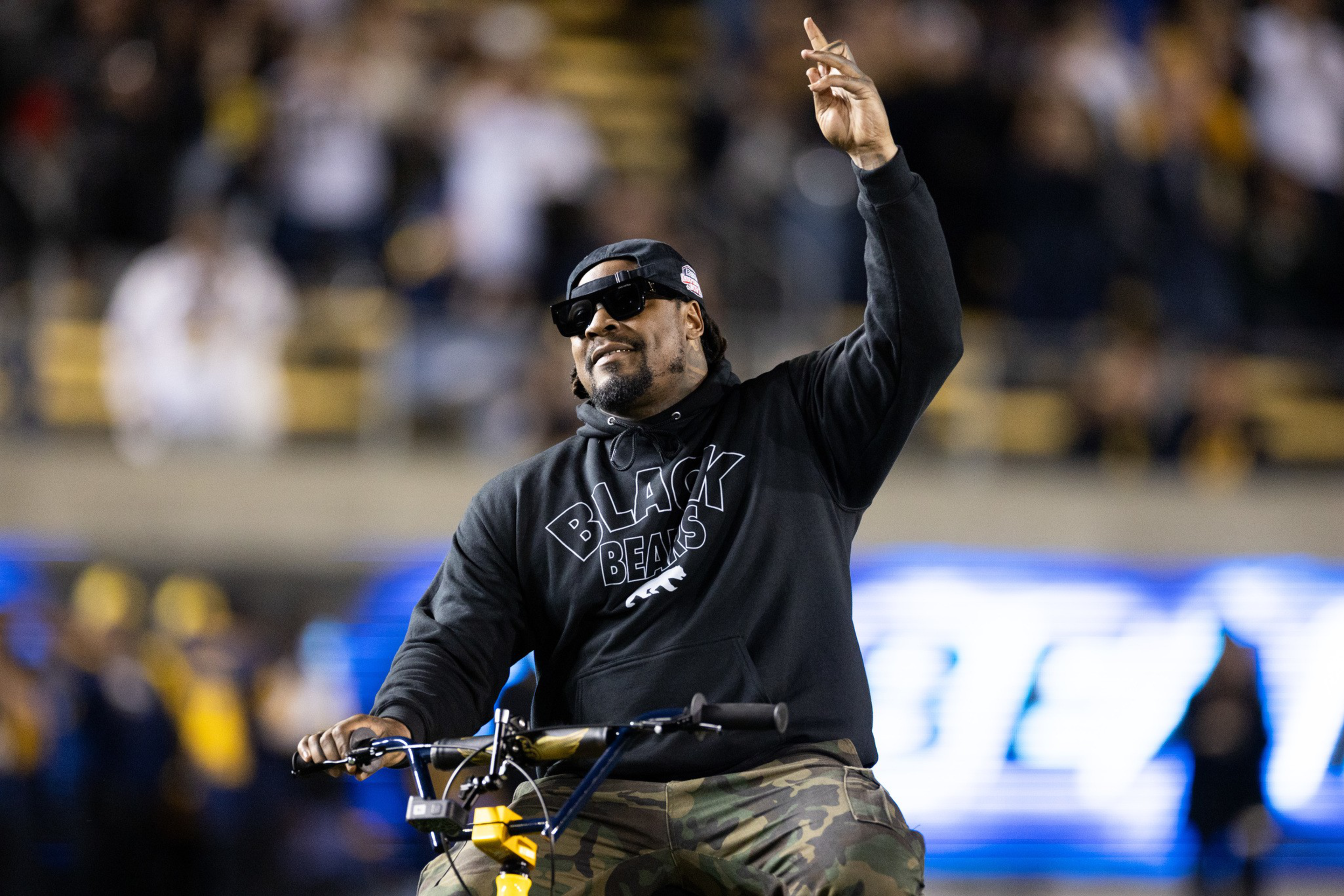A man wearing a black “Black Bears” hoodie, sunglasses, and a backward cap rides a bike and raises his hand at a crowded outdoor event.