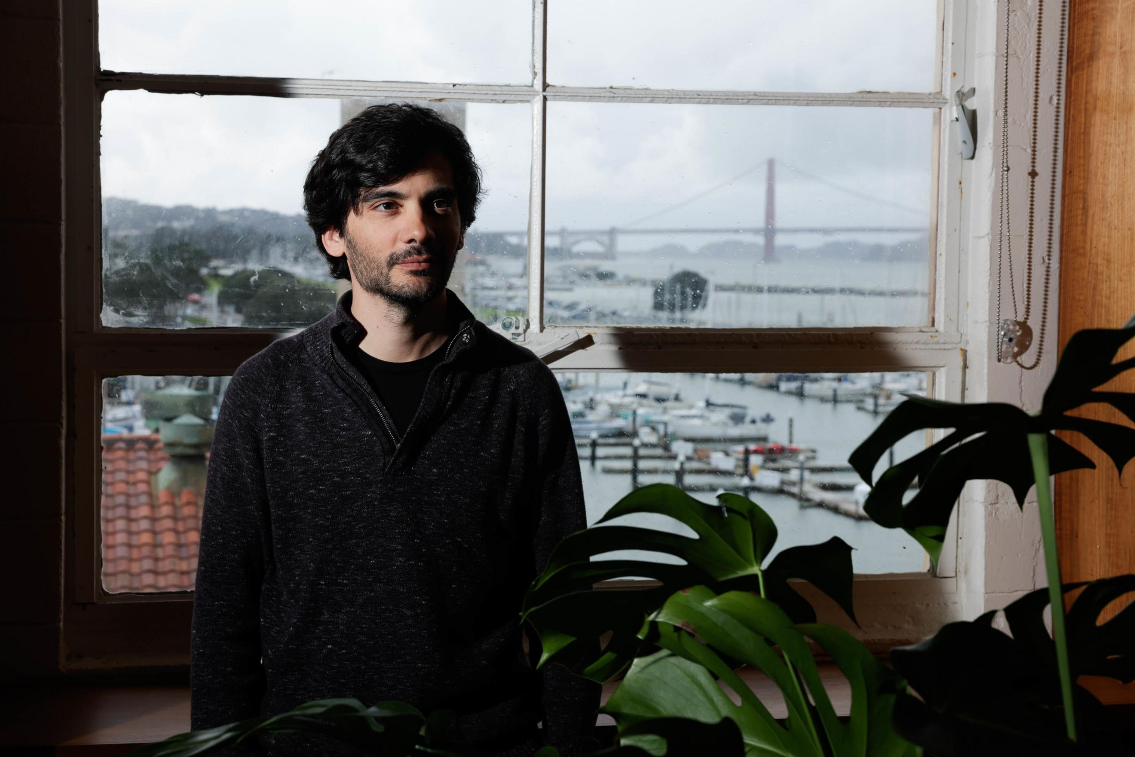 A man stands in front of a window with a bridge view, indoor plants, moody lighting.