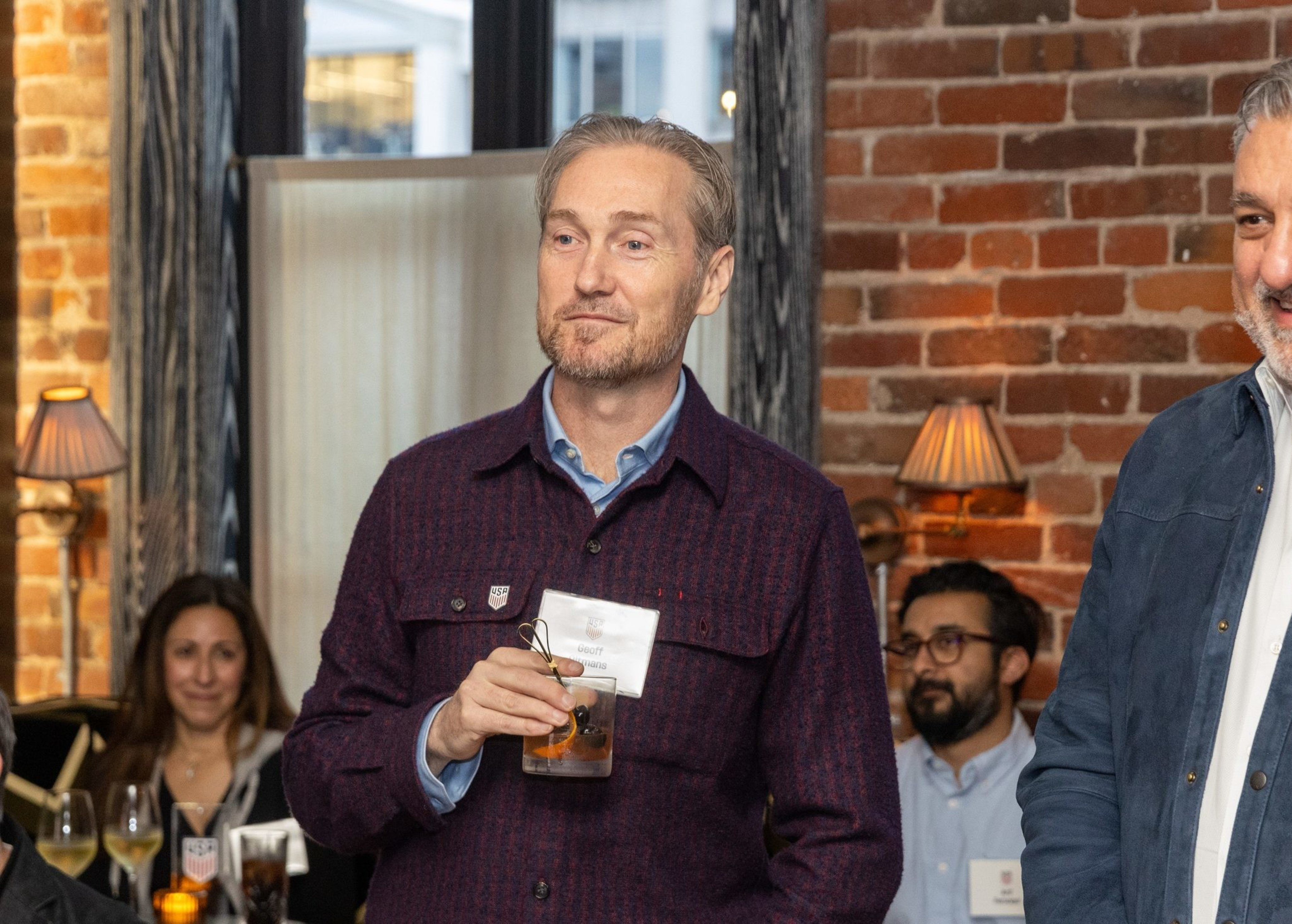 A man holds a drink and wears a nametag in a warmly-lit room with brick walls. Other people are seated nearby, and soft lighting adds a cozy atmosphere.
