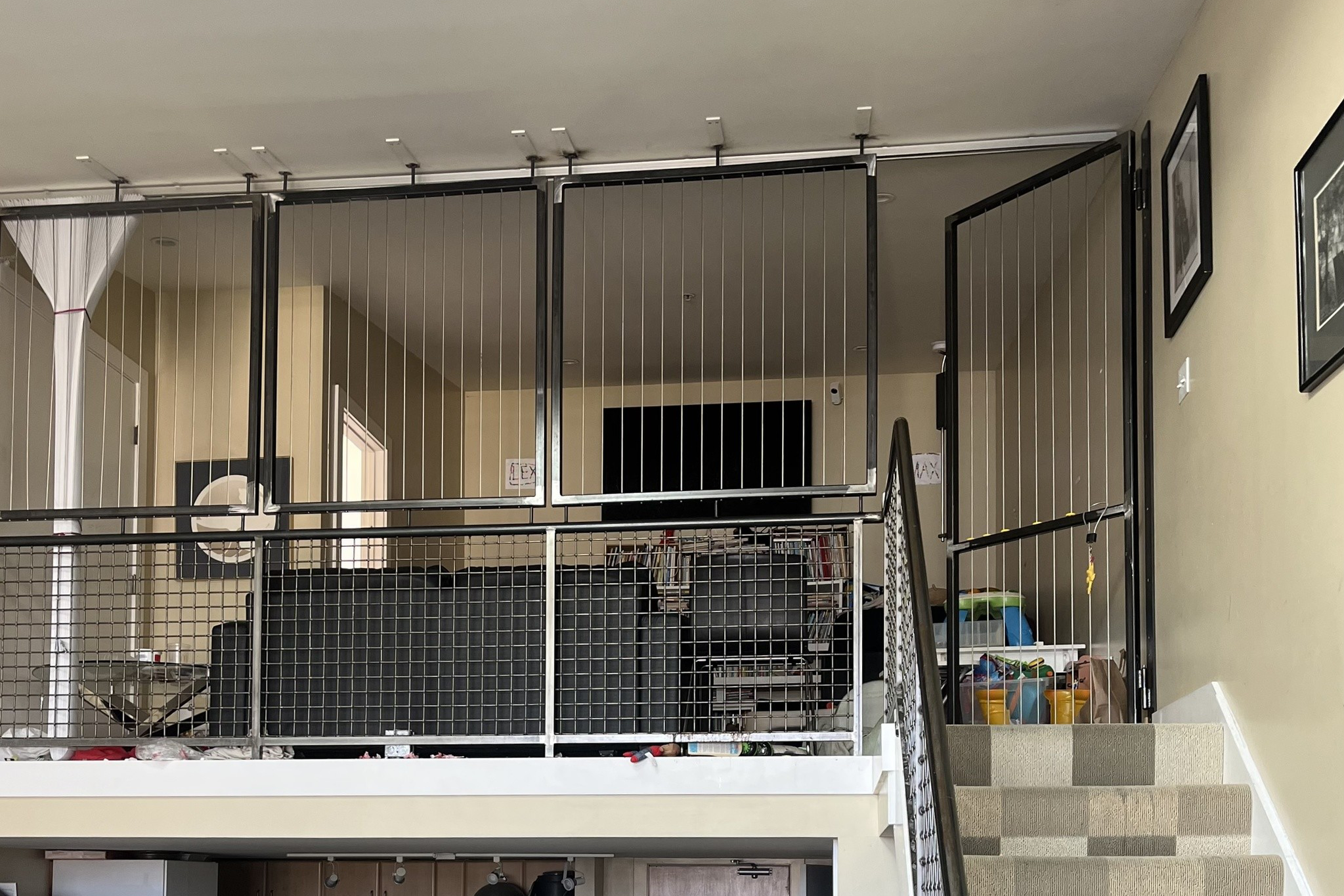 A loft area with black wire safety gates surrounds a living space with a TV and furniture, a staircase with patterned carpet leads up to it.