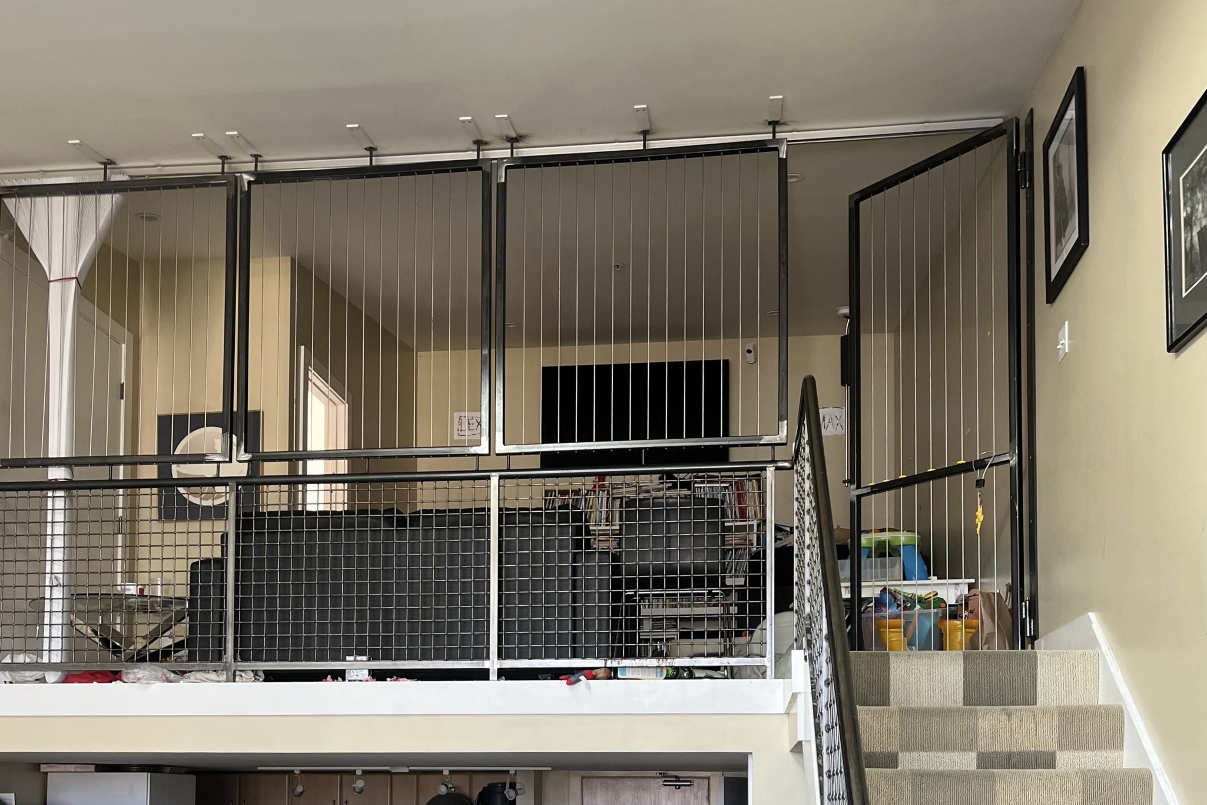 A loft area with black wire safety gates surrounds a living space with a TV and furniture, a staircase with patterned carpet leads up to it.